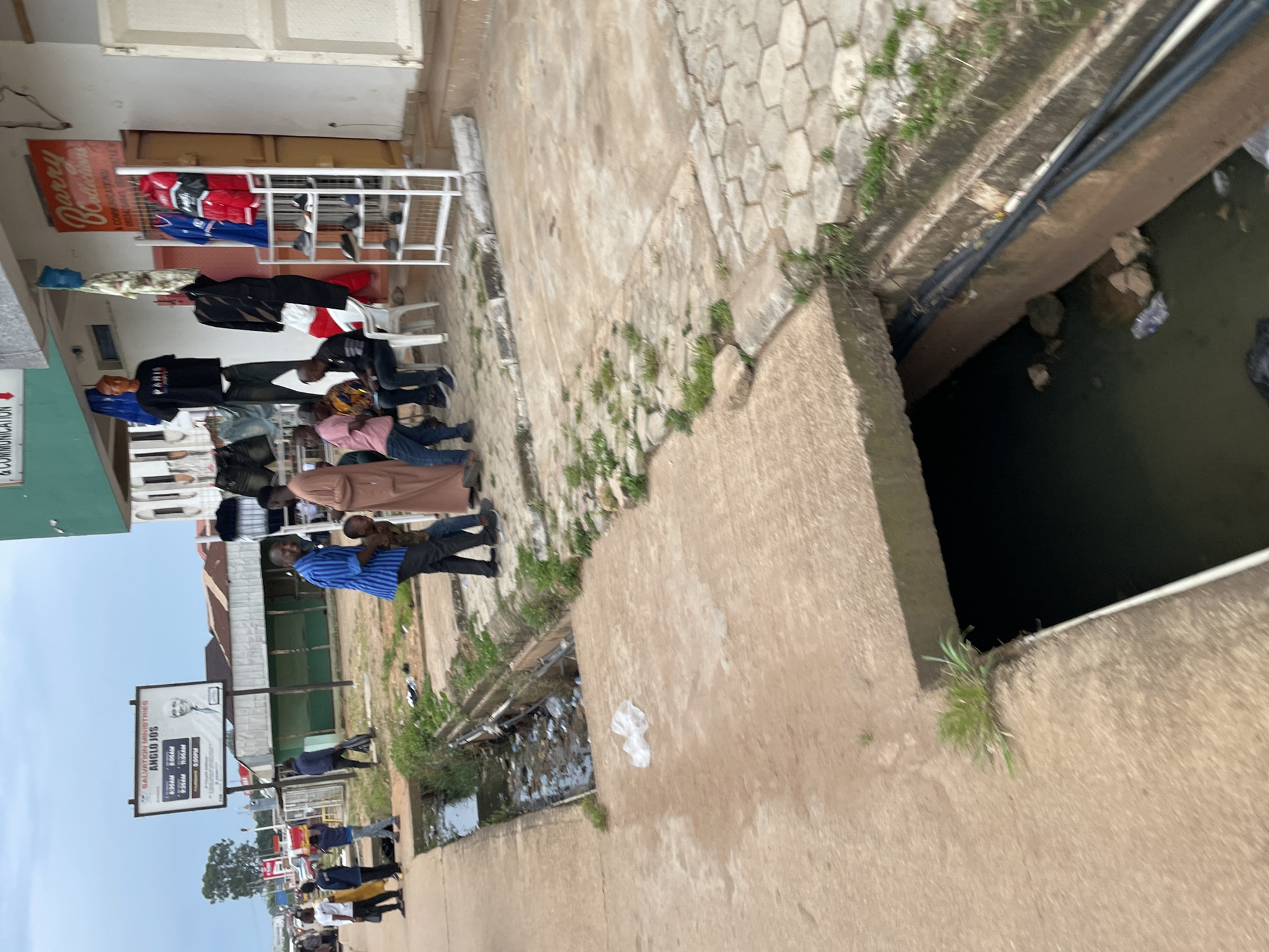 In Barakin Lerawi, Nigeria, a street scene unfolds. On the left, a group of people stand on a concrete sidewalk next to a drainage ditch filled with water and debris. Some are dressed in casual clothing, while others wear more formal attire. A man in a blue striped shirt smiles and interacts with a child. Further down the sidewalk, a sign announces "SALVATION MINISTRIES ANGLO JOS" with service times.
To the right, a shop displays clothing on racks and mannequins. Garments like t-shirts, shorts, and jackets are visible. A white shelf holds several pairs of shoes. The shop's facade features a sign that reads "Barry Collections." The overall environment appears to be an urban setting with buildings lining the street and an overcast sky above. The concrete sidewalk shows signs of wear and tear, and grass grows in patches along its edges. The drainage ditch is partially covered by concrete slabs, creating a channel for the water.