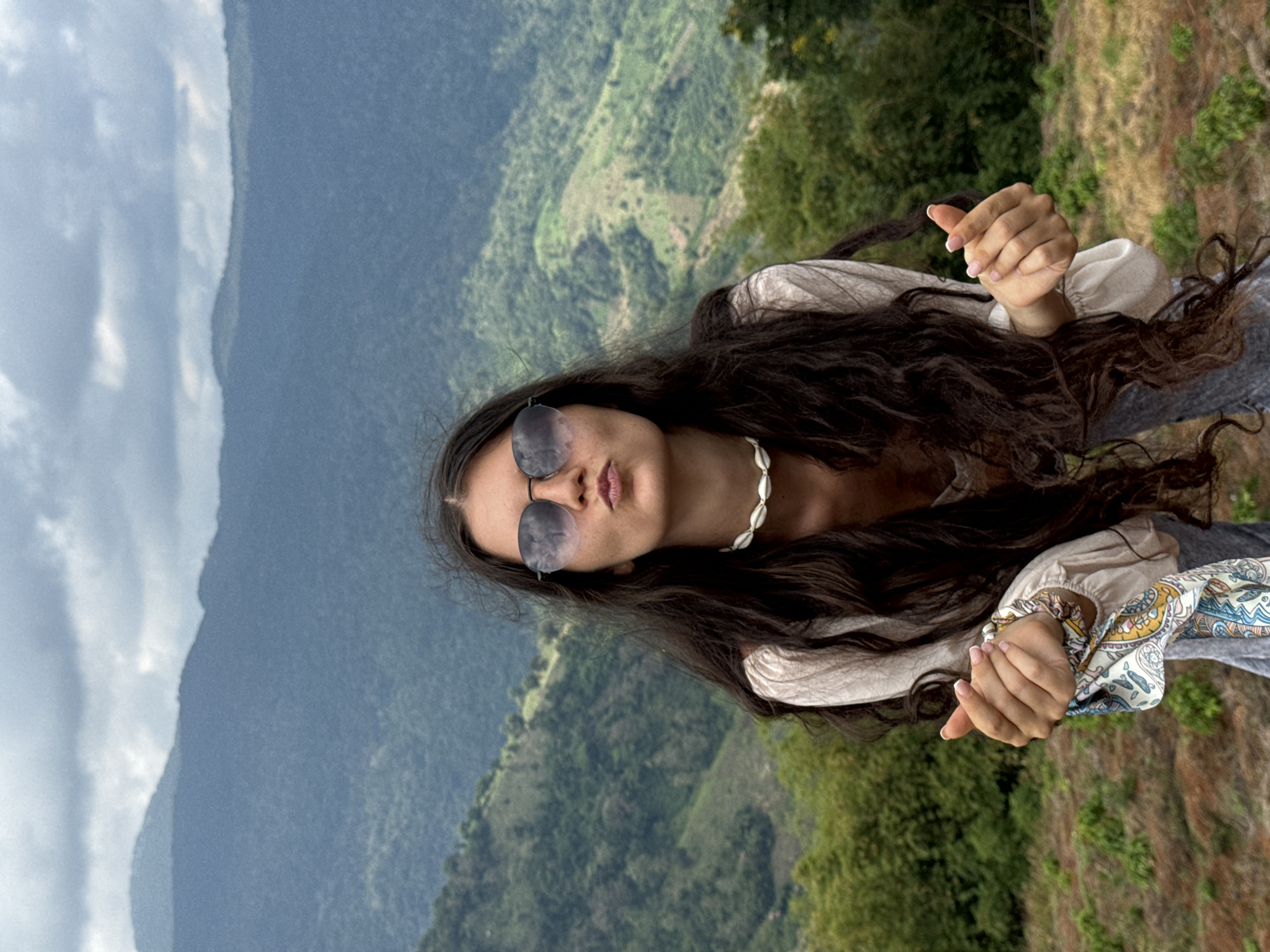 A young woman with long, dark, wavy hair and sunglasses is posing with her lips puckered in a kiss and her fingers forming a heart shape. She is wearing a light-colored blouse, a shell necklace, and gray pants. The background is a lush, green mountain landscape with some visible terracing and a cloudy sky. The image appears to have been taken outdoors. The specific location in Nao Sẻ, Vietnam, is not identifiable from the visual information provided in the image.