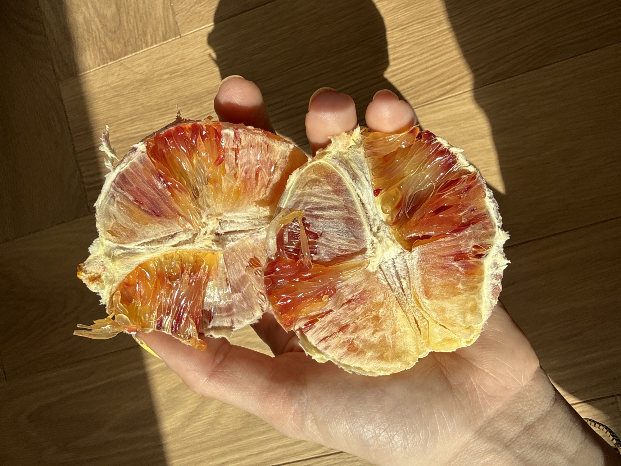 A hand holds a halved blood orange. The orange is partially peeled, revealing the red and orange flesh. The background is a wooden floor. The scene is lit with sunlight. The location is not identifiable as Moscow, Russia, from the provided image.