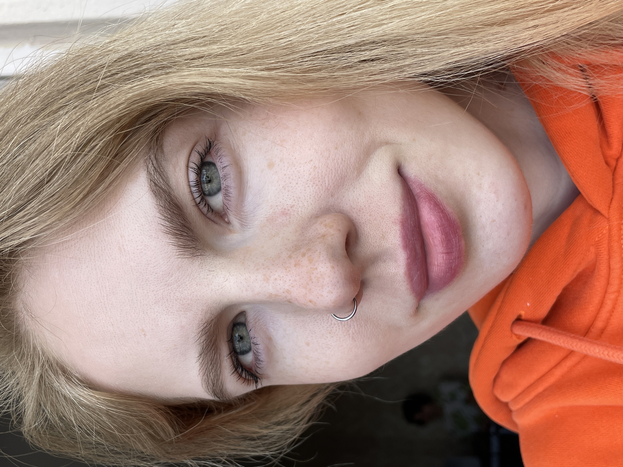 A close-up portrait captures a young woman from Izhevsk, Russia. She has long, light blonde hair and fair skin, noticeably adorned with freckles across her nose and cheeks. Her striking blue-green eyes, framed by full, light brown eyebrows and long eyelashes, engage directly or slightly off-camera. She sports a silver-colored septum ring in her nose. Her lips are painted with a matte pink or mauve lipstick, and she has a subtle, soft smile. The woman is wearing a vibrant orange hooded sweatshirt, with details of the fabric and drawstring visible. The lighting is bright and appears natural, suggesting a daytime setting. The image is rotated 90 degrees counter-clockwise. The background is indistinct and out of focus, offering no specific location cues beyond the subject herself. There is no visible text in the image.