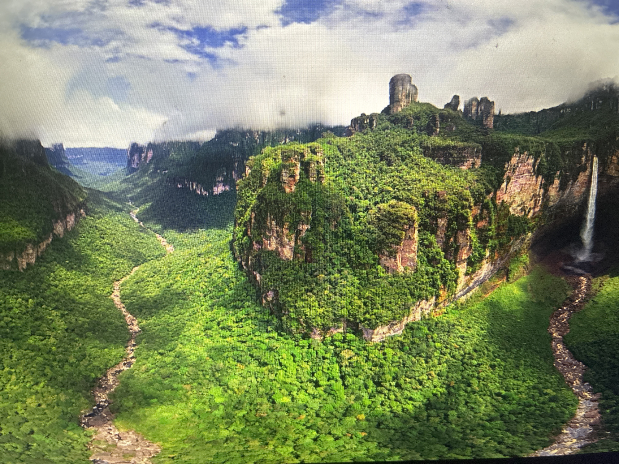 A wide aerial view captures a breathtaking, lush green mountainous landscape under a partially cloudy sky. The scene is dominated by massive, flat-topped mountains, or tepuis, that rise dramatically from the dense forest, their steep rock faces partially visible through the thick vegetation. Clouds hang low, occasionally obscuring the peaks and creating a misty ambiance.
Deep, verdant valleys cut through the terrain, with two distinct, winding rivers or streams snaking their way across the green expanse – one prominently in the foreground, and another receding into the distance on the left. A powerful waterfall cascades down a sheer cliff face on the right side of the image, feeding into one of the waterways below. The entire landscape is covered in an incredibly dense, vibrant green forest, suggesting a tropical or subtropical environment.
The lighting indicates daytime, with bright sunlight illuminating parts of the scene despite the cloud cover. The image conveys a sense of untouched wilderness and natural grandeur. No human presence or artificial structures are visible.