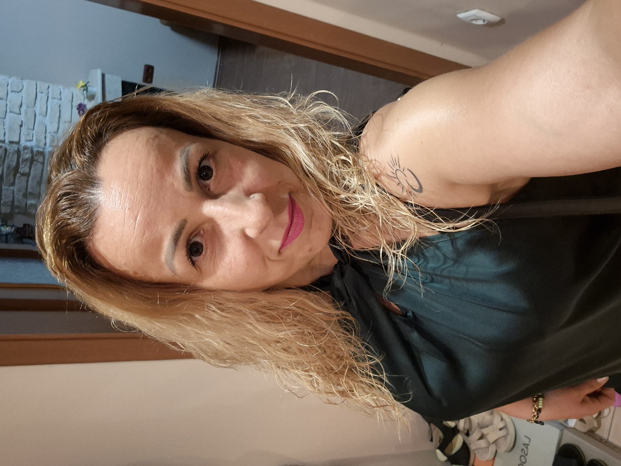 The image shows a blonde woman with wavy hair, wearing a dark green top. She has a nose ring and a tattoo of a sun on her left shoulder. She is standing indoors, in what appears to be a hallway or entryway, with a brick wall and a fireplace visible in the background. A doorway is to the right of the frame. Shoes are visible on a shelf in the lower left corner. The lighting suggests it is evening or indoors.