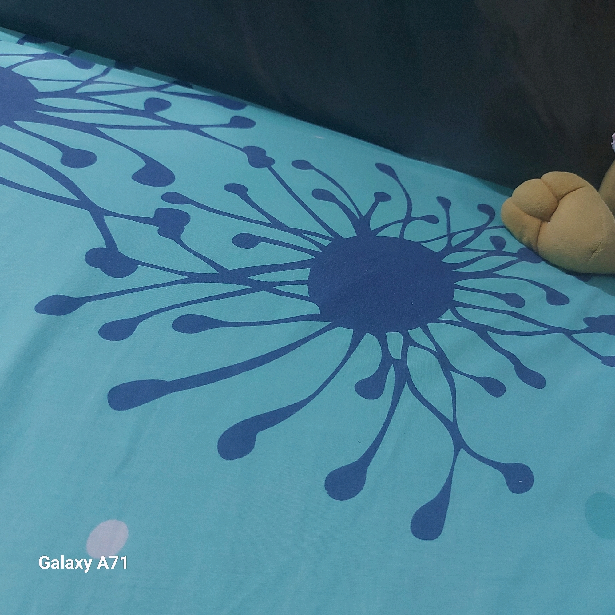 The image shows a close-up of a light blue fabric with a dark blue abstract design resembling dandelion seeds or splatters. A portion of a light yellow stuffed toy is visible in the upper right corner. The bottom left corner of the image contains a watermark that reads "Galaxy A71." The image's perspective is angled downwards, and the lighting suggests an indoor setting. There is no indication of the image being taken in Surabaya, Indonesia.
