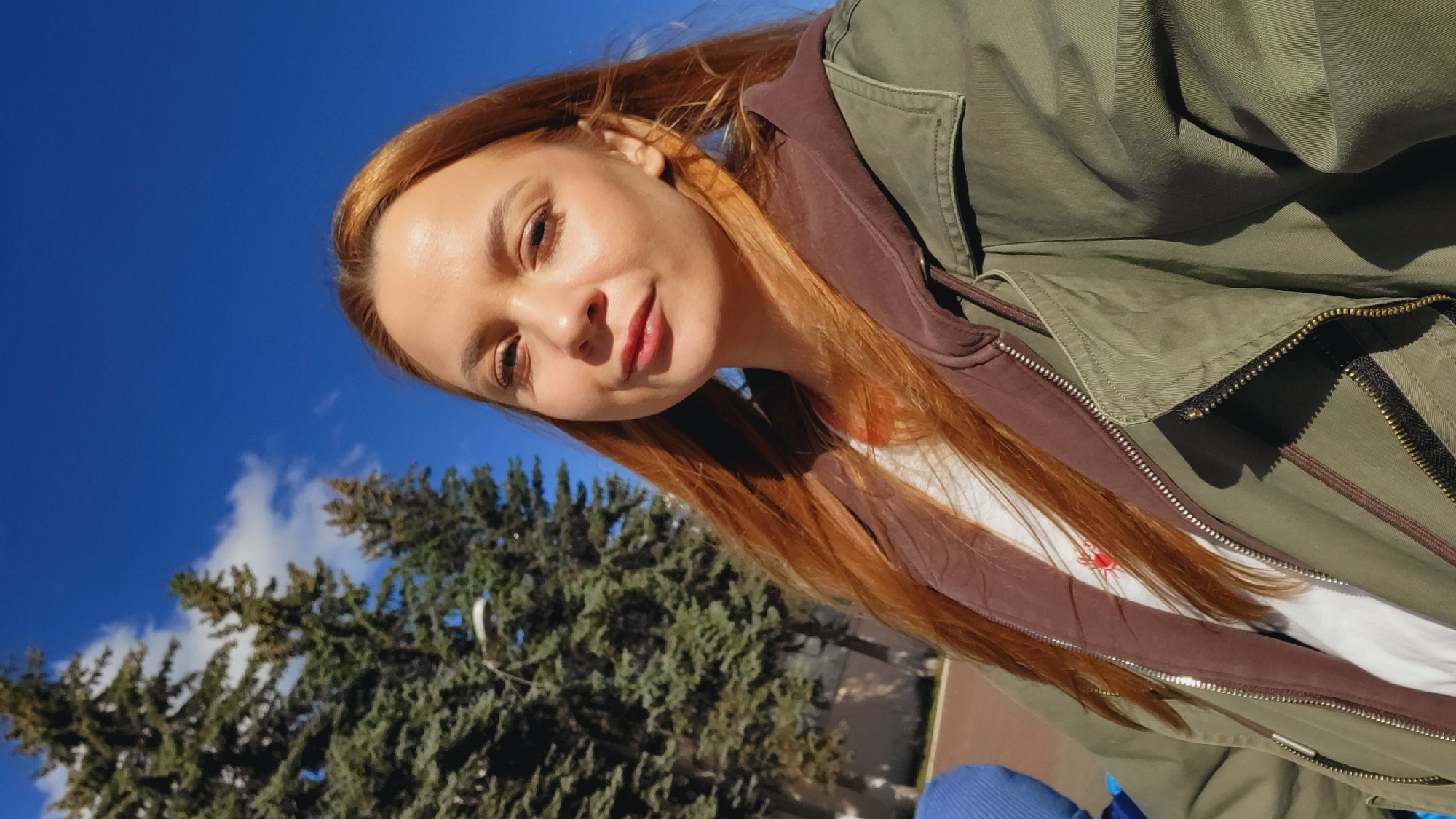 A young woman with long auburn hair, fair skin, and light makeup is seen in a selfie shot from a low angle. She is wearing a green military-style jacket over a brown hooded sweatshirt and a white t-shirt. The jacket is partially zipped up. The background consists of a bright blue sky with scattered white clouds and the top of a large, green coniferous tree.
Based on the limited visual information and a reverse image search, it is possible that this photo was taken in Chaikovsky, Russia, or a similar location with comparable urban greenery. However, without additional contextual details, it's impossible to confirm with certainty.