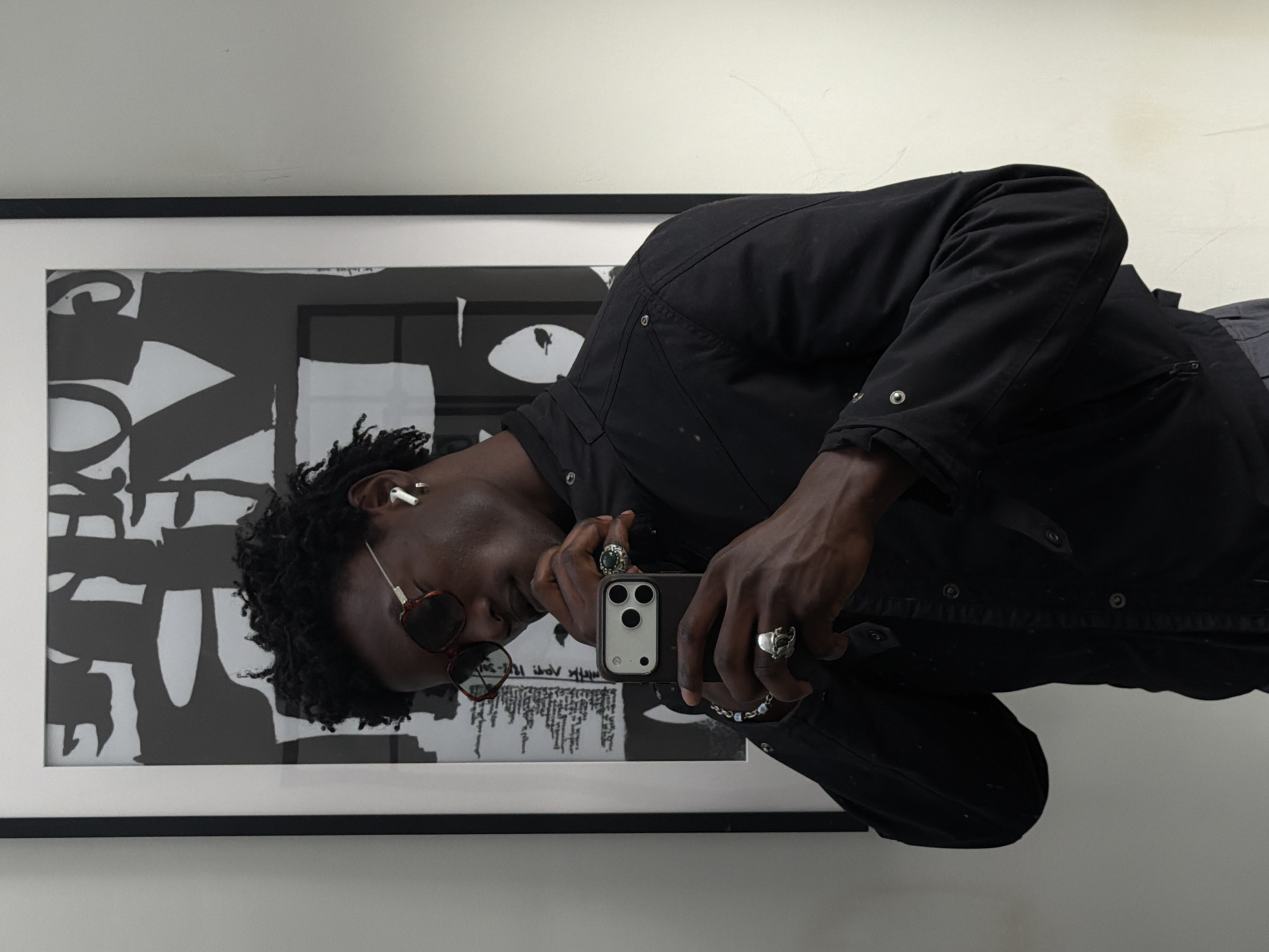 A person of color, wearing a black jacket, sunglasses, and an earpiece, is taking a selfie in front of a framed black-and-white abstract print. The individual is also wearing multiple rings. The location appears to be indoors, against a plain white wall. There is no discernible evidence to indicate that this is in the city of London, United Kingdom.