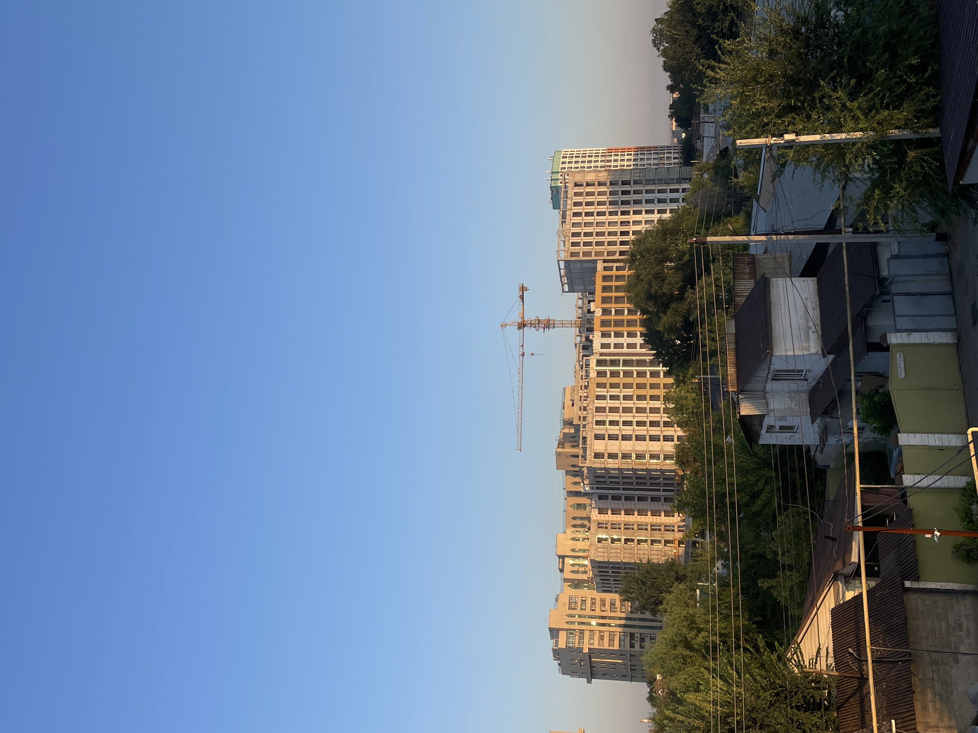 The image shows a construction site in the city of Almaty, Kazakhstan, under a clear blue sky during what appears to be sunset or sunrise. The foreground features a collection of low-rise residential buildings with corrugated metal roofs and some brickwork, interspersed with lush green trees and electrical wires. In the mid-ground and background, several multi-story apartment buildings are under construction, with one building displaying a facade of light brown and white panels, and another appearing to be a concrete structure. A large yellow construction crane is visible above the buildings, indicating ongoing development. The overall scene depicts a blend of older, established residential areas and new, modern construction projects in a semi-urban environment.