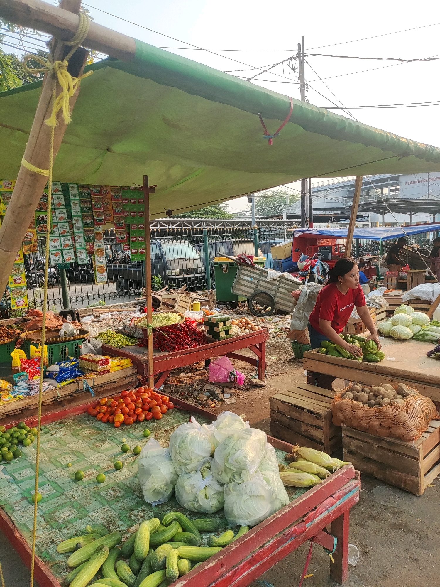 A woman in a red t-shirt is selling vegetables at an outdoor market stall in East Jakarta, Indonesia. The stall is covered by a green tarp supported by wooden poles and rope. Various vegetables are displayed on tables and in baskets, including tomatoes, cucumbers, limes, cabbage, corn, potatoes, and red chilies. Packaged snacks are also visible on shelves. In the background, a street scene shows other market stalls, vehicles, and a building with the sign "STASIUN KEBAYORAN". The ground is covered in dirt and debris.