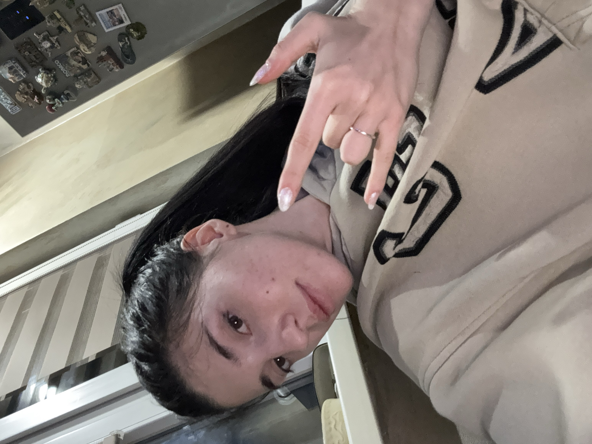 A woman with dark hair is posing for a selfie indoors. She is wearing a light-colored hoodie with black designs. She has a ring on her finger and is making a "rock on" hand gesture. Behind her, a window with blinds is visible. A wall with refrigerator magnets is also visible. The location cannot be determined from the image.