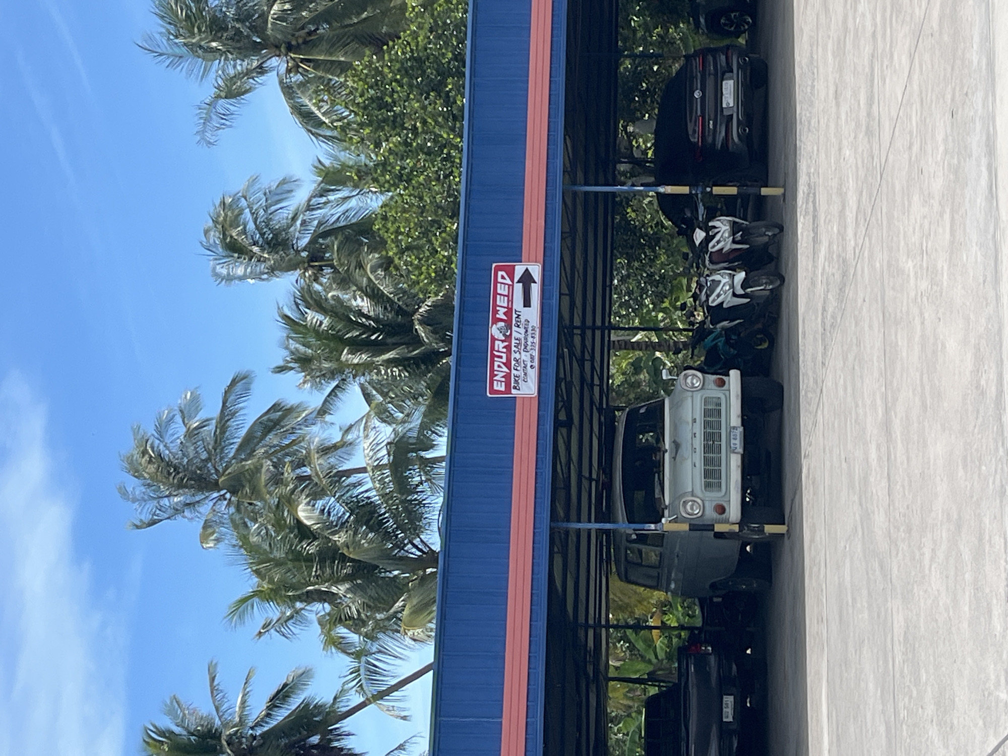 The image shows a parking area in front of a building with a blue and red facade in ตำบลเกาะพะงัน, Thailand. A sign on the building reads "ENDUROWEED BIKE FOR SALE / RENT CONTACT ENDUROWEED 087-335-8330" with an arrow pointing to the right. Several vehicles are parked under a covered area, including a light-colored vintage van, a black pickup truck, two motorcycles, and a dark-colored SUV. The background features lush green trees and palm trees under a clear blue sky.