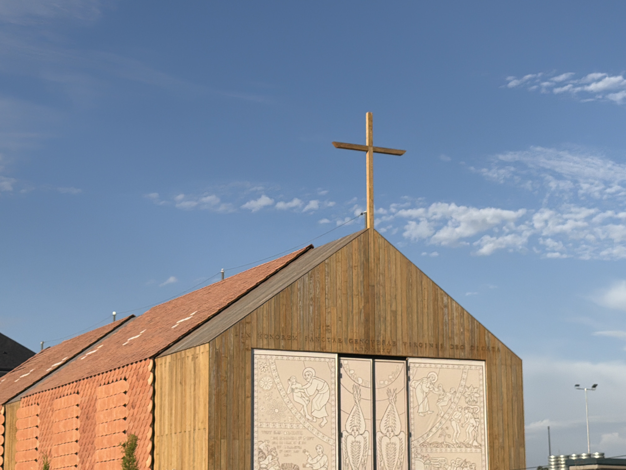 The image shows the exterior of a modern church in Majadahonda, Spain. The church features a distinctive architectural design with a prominent wooden facade and a sloping roof covered in red tiles. A large wooden cross stands at the peak of the roof. The facade is adorned with decorative panels depicting religious scenes in an artistic style. The sky is blue with scattered white clouds. The overall scene is one of contemporary religious architecture against a bright sky, suggesting a clear day.