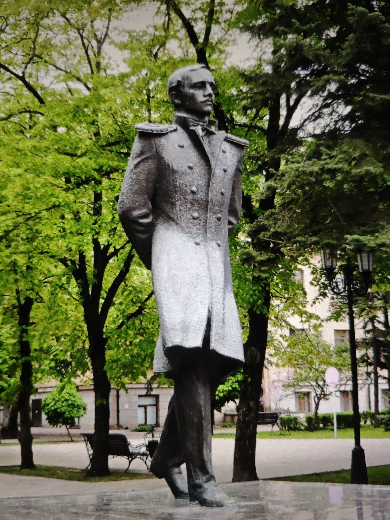 The image shows a full-body, dark-colored statue of a man in a military uniform. The man is depicted from the waist down and is looking to his left. He is standing in a park-like setting with lush green trees in the background. There are buildings with windows and a lamppost visible behind the statue. The ground in front of the statue appears to be wet, possibly from rain, and is reflective. In the background, there are also benches and a pink circular sign. This statue is located in Stavropol, Russia, specifically in the Alexander Garden.