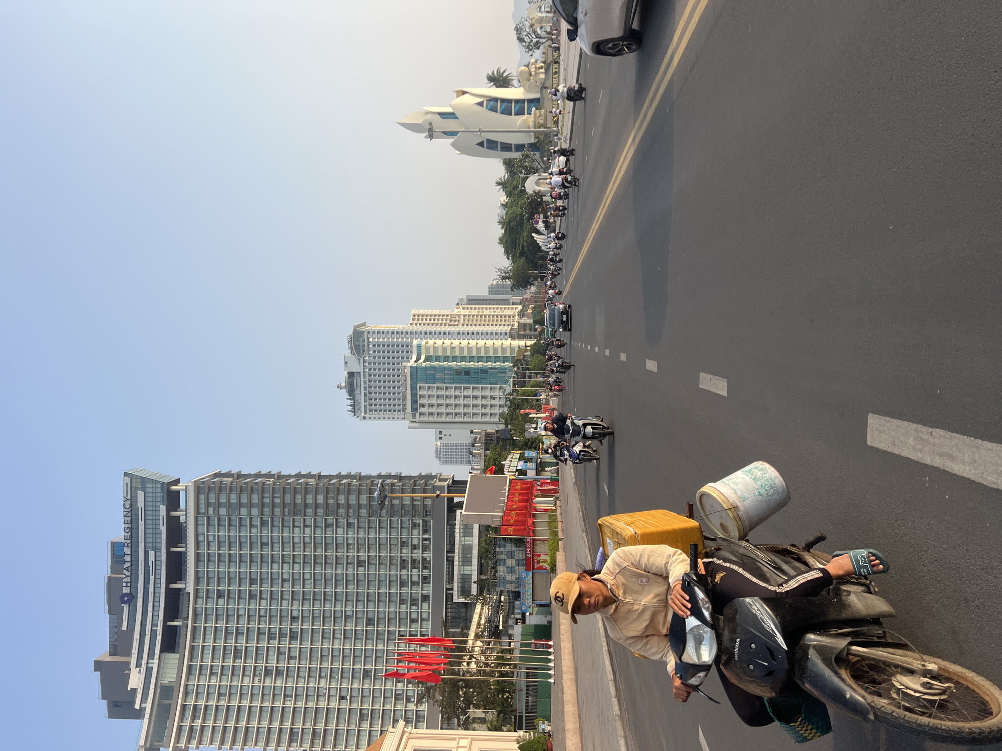 The image shows a street scene in Nha Trang, Vietnam. A man on a motorcycle is in the foreground, with a large, yellow package and a white bucket strapped to the back. The street is busy with traffic, primarily motorcycles, heading away from the camera. Tall buildings are visible on the left, including what appears to be the Hyatt Regency Nha Trang. Red flags line the street in front of the buildings. In the distance, a white building complex can be seen on the right, near the beach. The sky is hazy and blue.