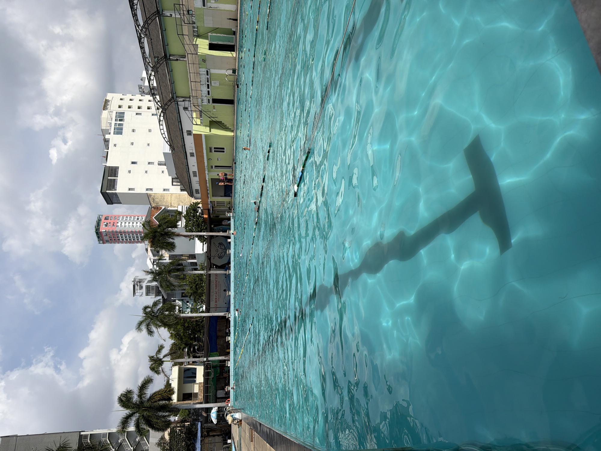The image depicts a swimming pool in Nha Trang Ward, Vietnam, under a partly cloudy sky. Adjacent to the pool are buildings, including a white apartment complex and structures with green accents. Palm trees are interspersed between the buildings and the pool area. The pool water is clear and reflects sunlight, creating ripples and shadows. The pool is divided into lanes by ropes and buoys. A long shadow of a T-shaped object is visible in the water. Several people are at the edge of the pool, with some appearing to be preparing to swim.