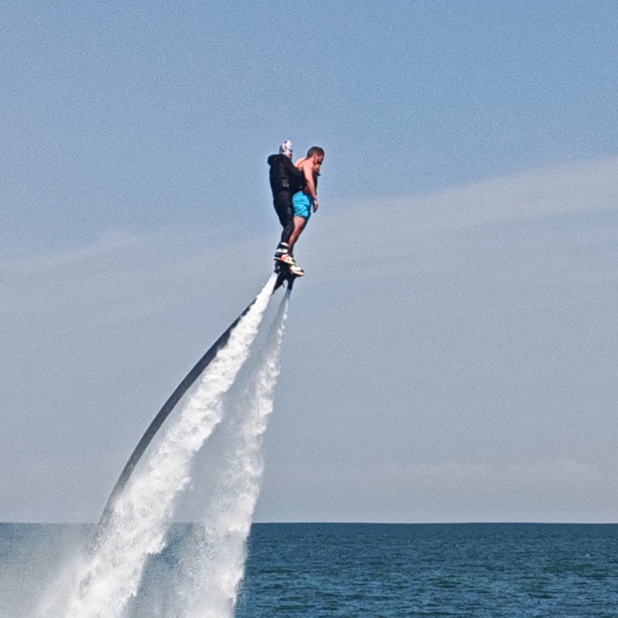 In Unknown, Unknown, two males are airborne above a body of water, supported by a flyboard. The lead individual, wearing a black wetsuit and a white-and-black patterned helmet, has their feet strapped to the flyboard, which expels two powerful white water jets downwards. A dark hose connects the flyboard to the water surface. A second male, shirtless and in blue swim trunks, is piggybacking the lead, with his left arm around the lead's neck. He has a beard and sunglasses on his head. The sky is clear and light blue, meeting a calm, deep blue-green sea at the horizon. The water below the flyboard is disturbed by the propulsion.
