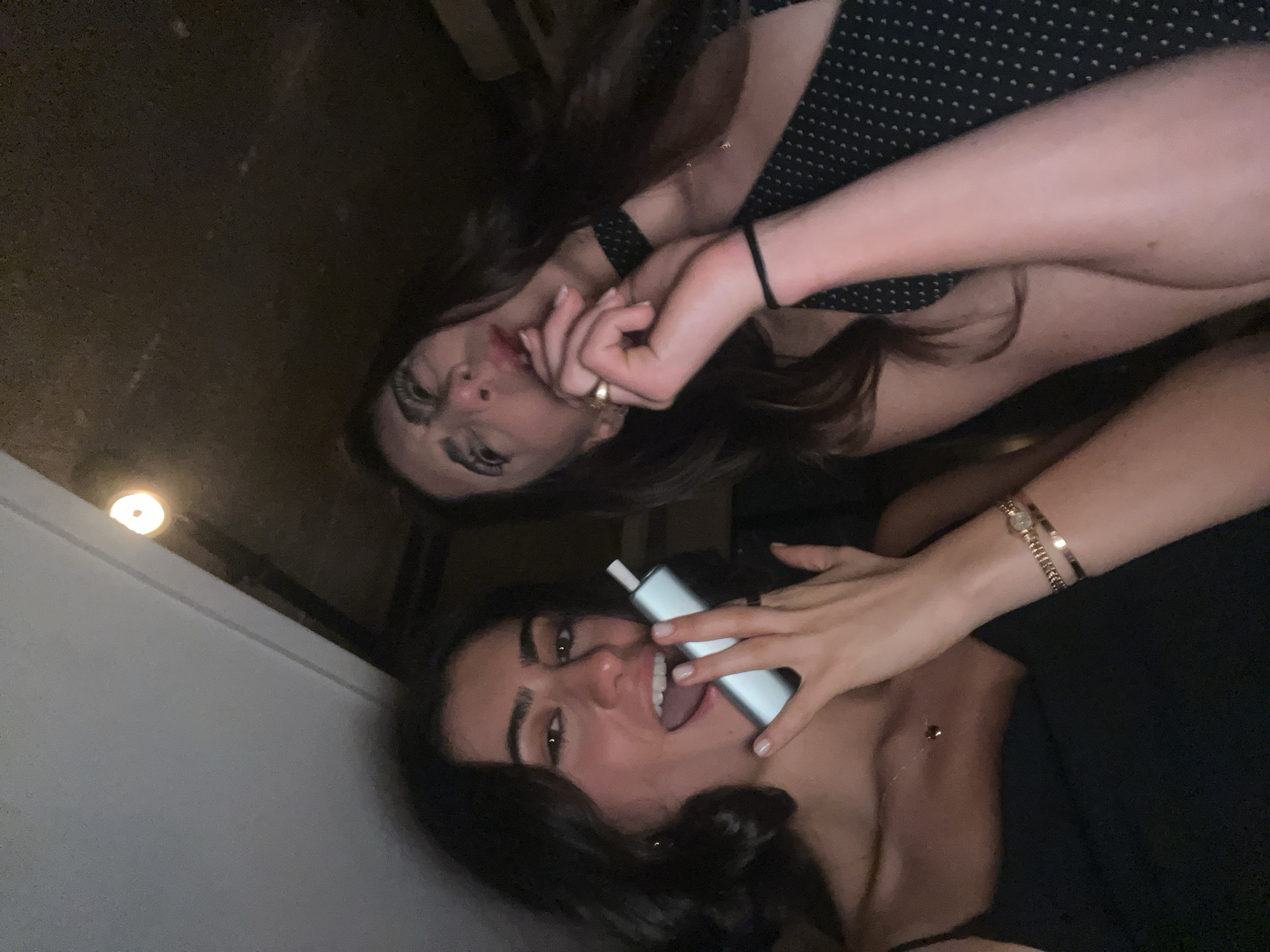 Two women are lying down, seemingly in an indoor setting. The woman on the bottom is holding a white, rectangular object, possibly a vape or electronic device, near her mouth with her tongue slightly protruding. She has dark hair, light skin, and is wearing a dark top. The woman above her has brown hair and light skin, and is wearing a black and white polka dot top. She has her hand near her face. A door and ceiling light are visible in the background. There is no verifiable information to indicate the image was taken in Paris, France.