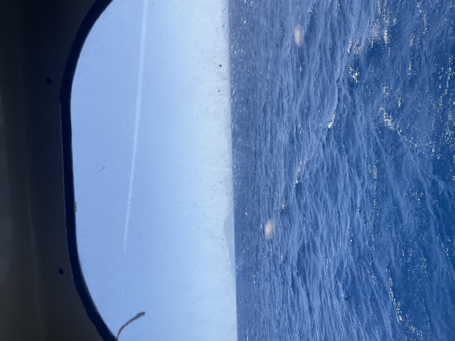 The image displays an expansive view of the open sea and sky, observed through an arched window. The foreground features deep blue, moderately agitated ocean water, illuminated by direct sunlight creating numerous shimmering reflections on its surface. A low, hazy landmass, possibly distant islands or coastline, is visible along the horizon. The sky above is clear and light blue, traversed by a single, prominent white aircraft contrail positioned horizontally across the upper third of the frame. The windowpane itself exhibits several small, diffuse light artifacts and minor surface imperfections. This maritime scene is observed from the sea in the vicinity of Marsala, Italy. No human subjects, specific vessels, or additional objects are identifiable within the primary view beyond the natural elements and the viewing portal.