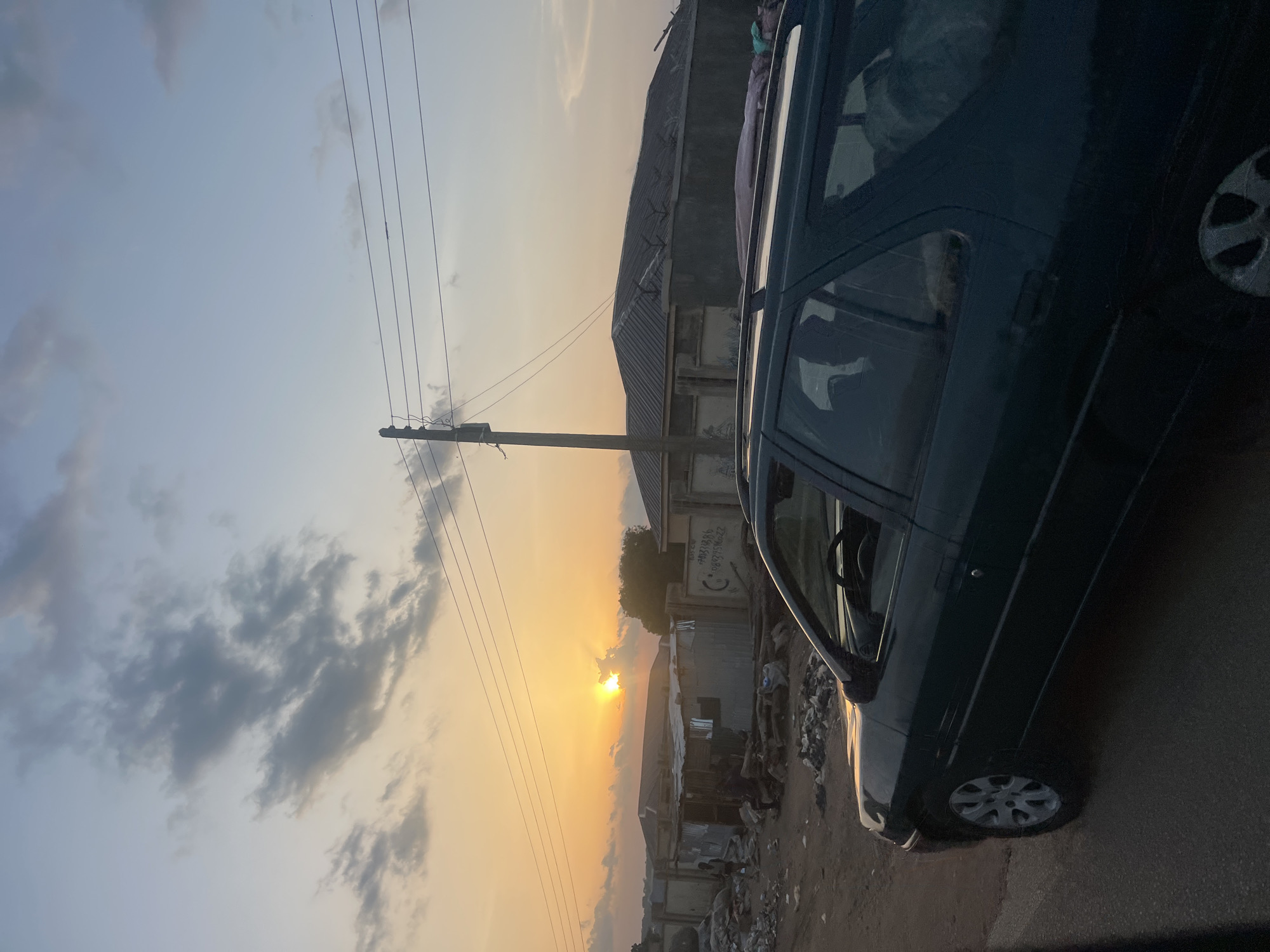 The image depicts an outdoor scene at sunset or sunrise in Rinjim Gani, Nigeria, characterized by warm light from a low sun. The sky transitions from a bright orange-yellow near the horizon, where the sun is partially obscured by clouds, to lighter blues and grays at the top, with scattered cumulus clouds present.
In the foreground, a dark-colored Peugeot 306 Break (station wagon) is positioned on the right, with its passenger side visible. The vehicle appears to be on a paved or hardened dirt surface. A light-colored item, possibly a blanket or animal, is discernible through the rear passenger window. Reflections of the sun are visible on the car's bodywork.
The midground features a dirt road or open ground on the left, strewn with significant amounts of litter, including plastic bags and debris. Several single-story buildings are visible, some constructed with corrugated metal sheets, others with concrete walls and corrugated roofs. One concrete wall includes what appear to be handwritten phone numbers or codes: "0803713006" and "08092758022." This wall is topped with barbed wire fencing.
Multiple human figures are present in the midground, near the structures and litter. One figure, possibly a child, stands on the far left near the corrugated metal structures. Another figure is seated or crouched amidst the litter closer to the center. Their specific actions are indistinct due to distance and lighting.
A wooden utility pole with multiple horizontal power lines extends vertically through the mid-left portion of the frame, carrying lines across the sky. In the background, beyond the immediate structures, additional indistinct buildings and sparse vegetation are visible under the horizon line.