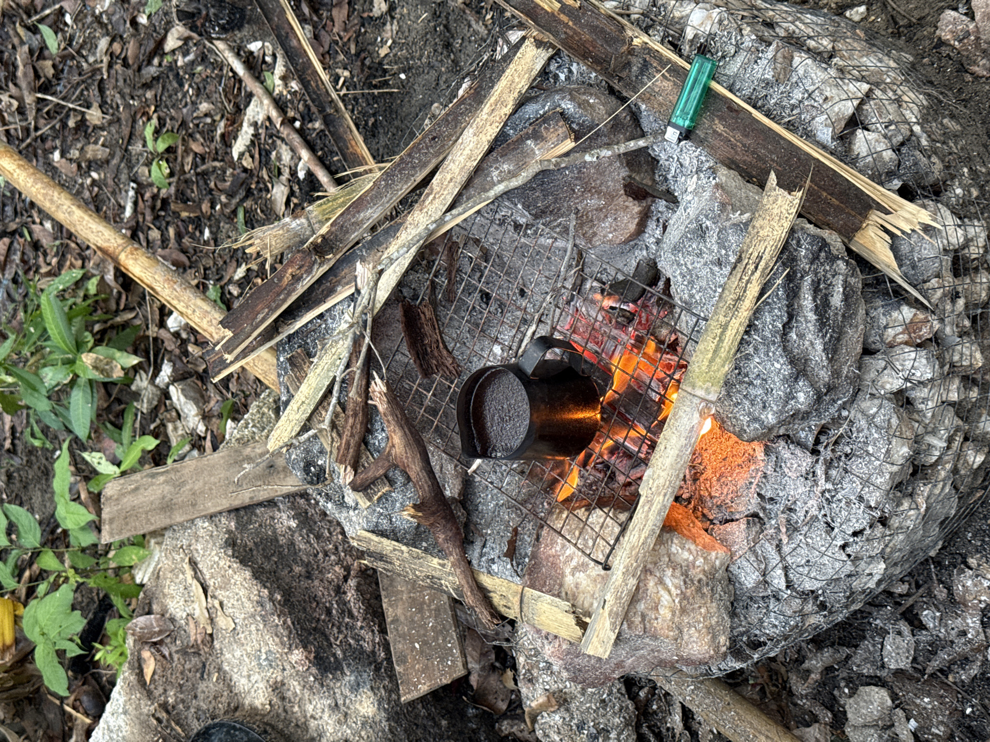 A black metal cup sits on a wire grill over a fire. The fire burns within a rock structure covered in wire mesh. Broken sticks and twigs are scattered around the fire pit. A green lighter rests on a rock next to the fire. Green plants grow to the left of the scene. The location is Moo 7, Thailand.