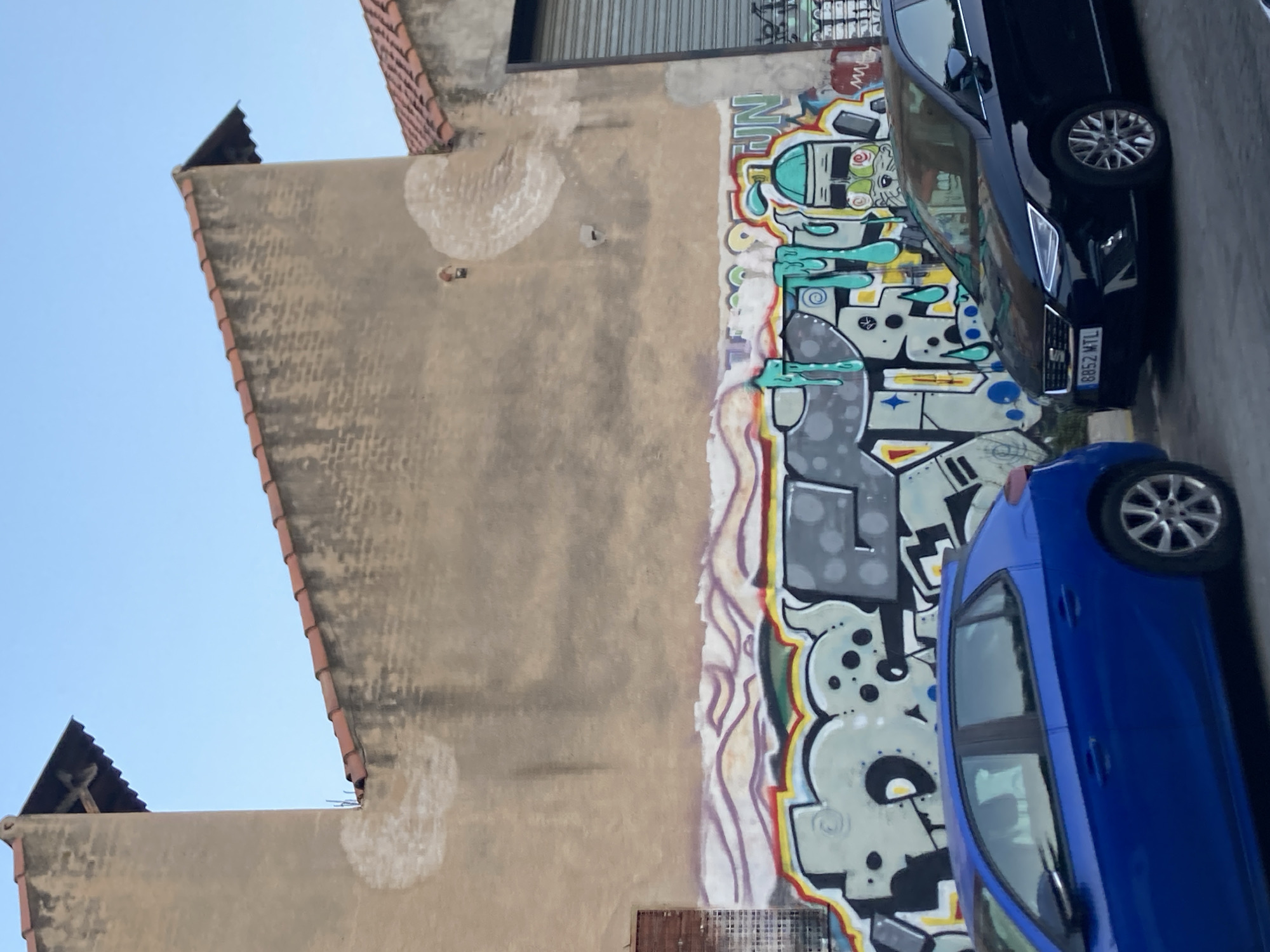 The image depicts a section of a building wall in Badalona, Spain, adorned with colorful graffiti. Two parked cars, one blue and one black, are in the foreground. The building's exterior wall is a light tan color, showing signs of weathering and some circular, lighter patches. The roofline, with terracotta tiles, is visible at the top of the frame, as is a section of a metal roller shutter on the right side. The graffiti covers the lower portion of the wall, featuring a variety of shapes, letters, and cartoon-like figures rendered in bright blues, greens, yellows, reds, and grays. The sky above is a clear, light blue. The black car's license plate reads "8852 MTL."