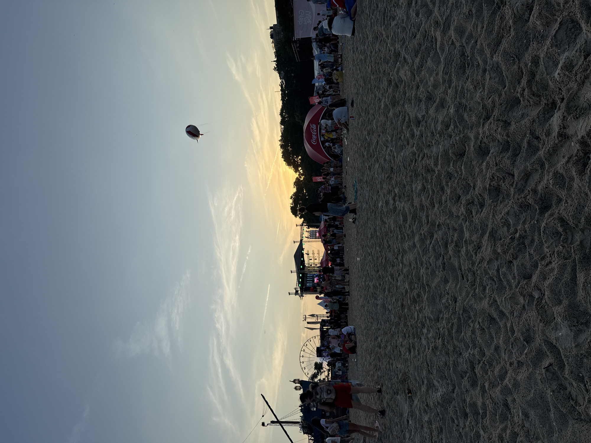 This is a photograph taken at the beach in Varna, Bulgaria, during a festival or event. The image captures a wide sandy beach filled with people gathered for an outdoor event. In the background, there are stages, tents, a Ferris wheel, and other structures associated with a festival. The sky is clear with some wispy clouds, and the sun appears to be setting, casting a warm glow over the scene. A blimp or balloon with red and white stripes is visible floating in the sky. The overall atmosphere suggests a lively and festive occasion.