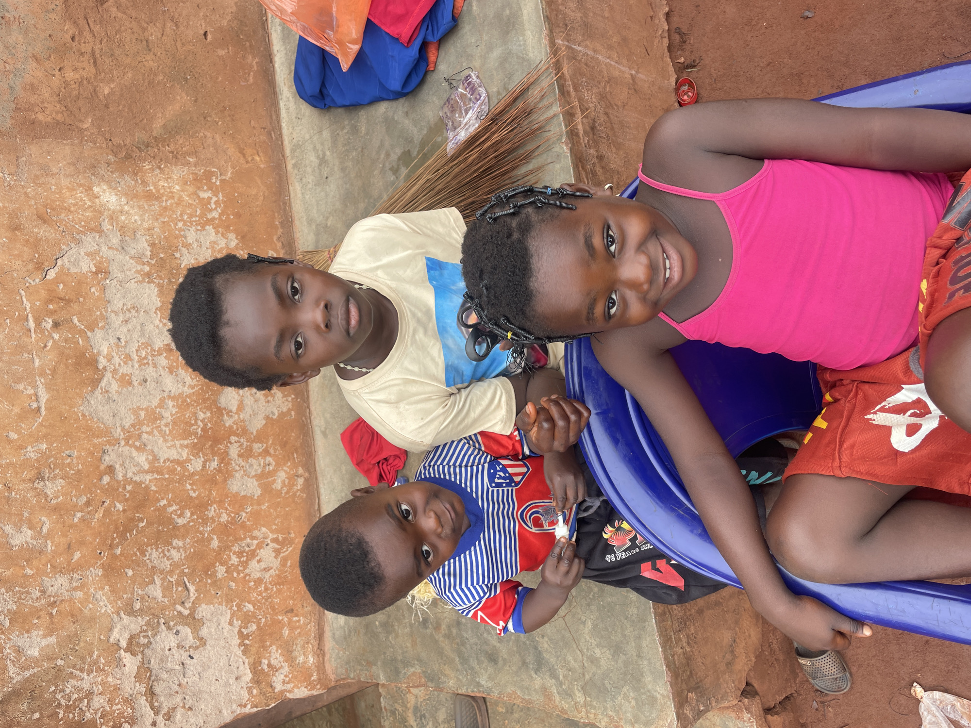 Three children are positioned close together, looking up towards the camera. The child on the right is in a pink tank top and red shorts and is seated inside a blue plastic object. The child in the middle is wearing a white shirt with a blue image, and the child on the left wears a vertically striped blue, white, and red shirt. The children appear to be located in the city of Ogbe, Nigeria. In the background, a broom, clothing, and debris are visible. The ground appears to be composed of reddish-brown earth with patches of concrete.