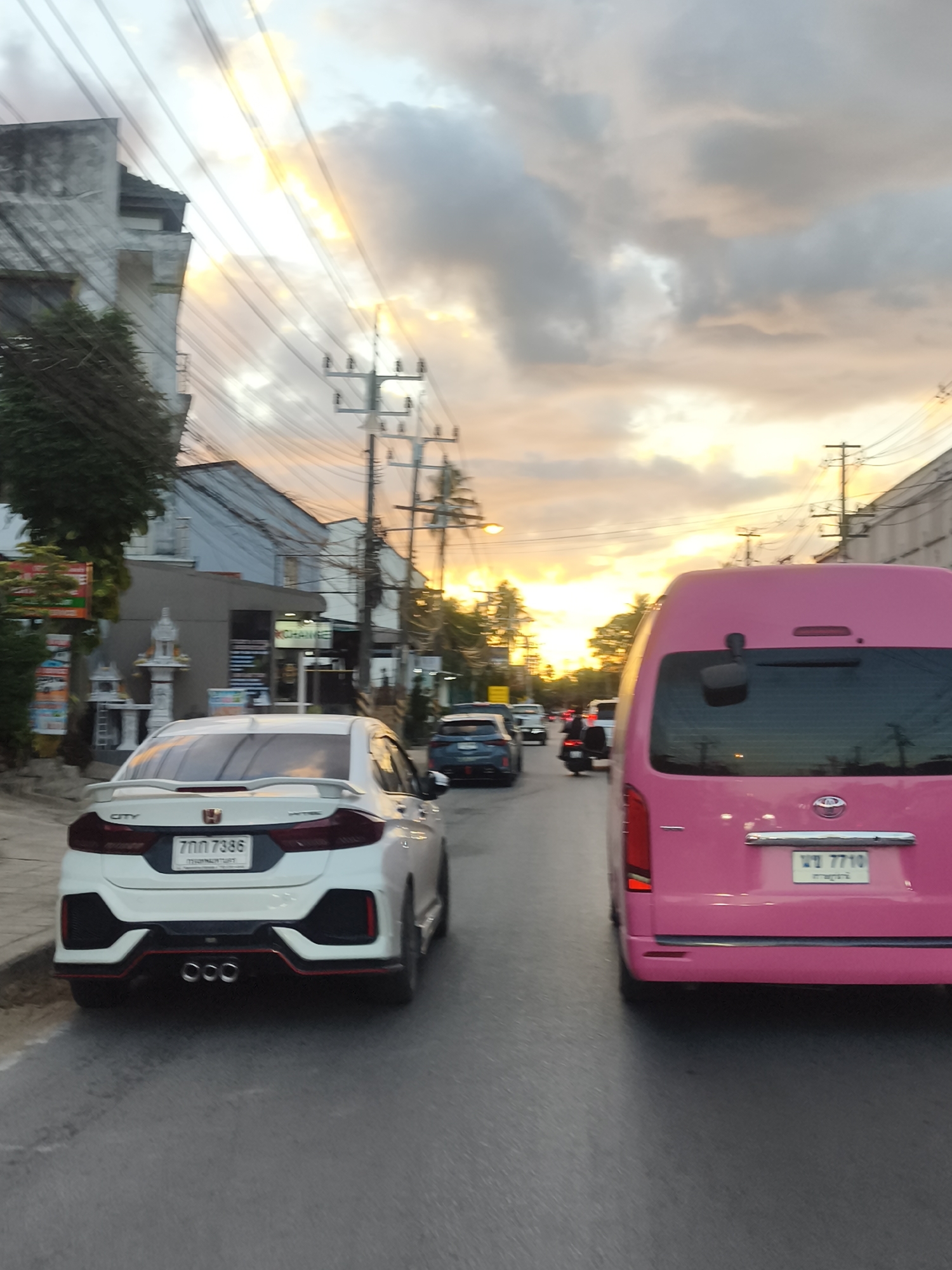The image depicts a street scene in Baan Bo Phut, Thailand, likely during a sunset or sunrise. The sky is filled with dramatic clouds illuminated by warm, golden light, suggesting the golden hour. Several vehicles are on the road, indicating a moderate level of traffic.
In the foreground, the rear of a white sedan is visible, featuring a sporty design with a spoiler and triple exhaust pipes. Its license plate reads "7กภ 7386" in Thai script. To the right, a bright pink van is prominently featured, also with its rear facing the camera. Its license plate displays "พ 7710" in Thai. Other vehicles, including a dark-colored SUV and a motorcycle, are present further down the road.
The roadside features buildings with a mix of architectural styles, including some older structures with weathered facades and others that appear more modern. Electrical poles and wires crisscross the scene, a common sight in many urban and semi-urban environments. On the left side of the road, there are small shrines or spirit houses adorned with offerings, a cultural detail often seen in Thailand. One building has signs with Thai text, one of which appears to say "CHANGE". The overall atmosphere is that of a typical evening on a busy street in a tropical location, with the natural beauty of the sky adding a striking visual element.