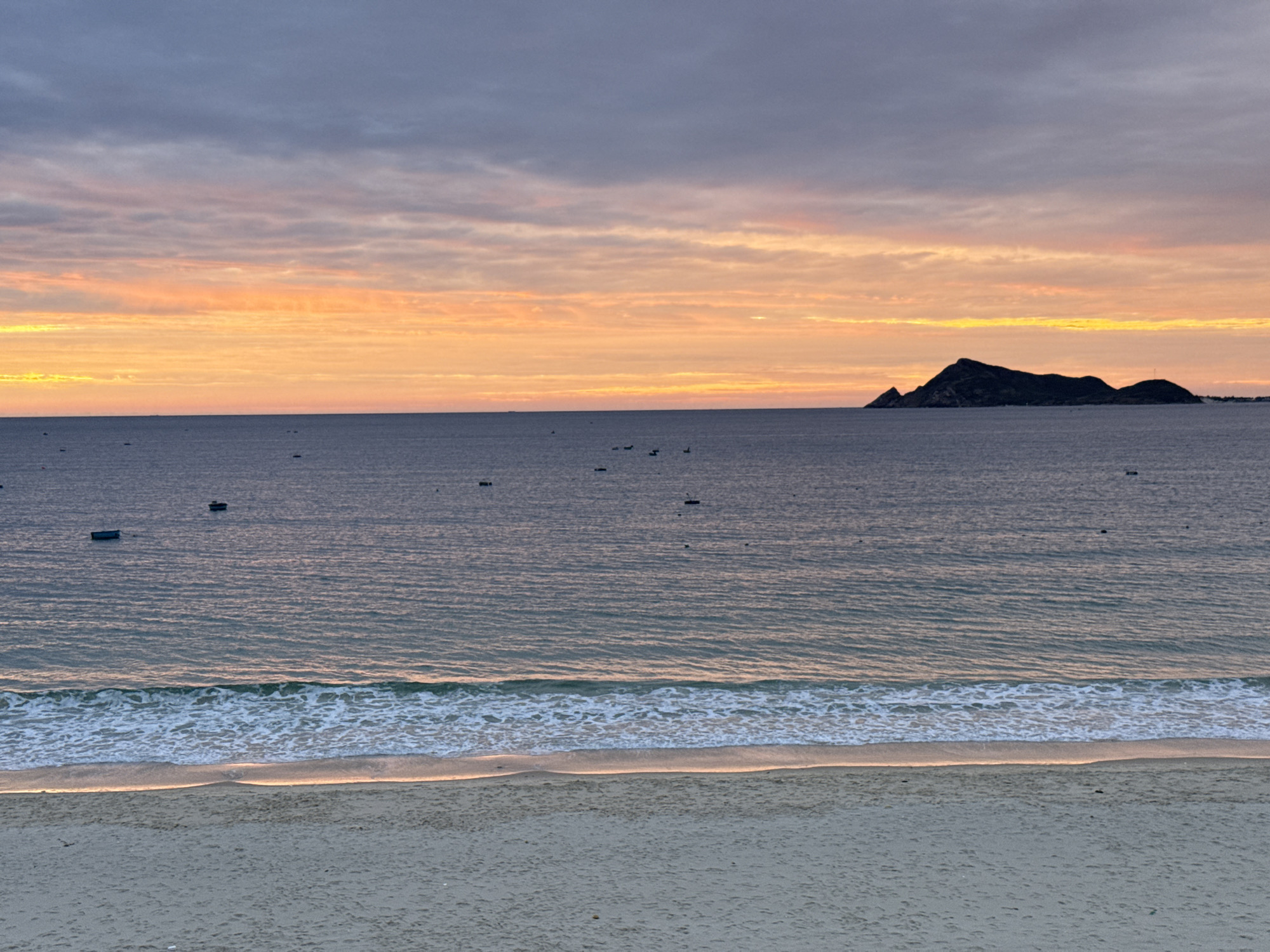 This is an eye-level, full shot of a serene seascape at what appears to be dawn or dusk. The scene is set at the beach in Song Cau, Vietnam, a coastal town known for its tranquil beaches. In the foreground, a wide expanse of pale sand stretches across the bottom of the frame, showing subtle textures and footprints. A gentle wave line breaks upon the shore, with foamy white crests and the faintest hint of golden light reflecting on the wet sand.
The middle ground is dominated by the vast, calm sea. The water is a muted gray-blue, with subtle ripples and a soft sheen from the low-angled light. A scattering of small, dark objects, likely fishing boats, are visible on the water's surface, some appearing as dark rectangles and others as more indistinct dots.
The horizon line is sharp and clear, separating the sea from the sky. The sky above is a dramatic display of color. The lower portion is a vibrant band of orange and yellow, indicative of sunrise or sunset, with soft, horizontal clouds. Higher up, the sky transitions to a dusky purple and gray, with darker, more textured clouds streaked across the expanse. In the distance, a dark, silhouetted island with a rugged peak rises from the sea, adding a focal point to the far right of the horizon. The overall mood is peaceful and tranquil, capturing the natural beauty of the Song Cau coastline.