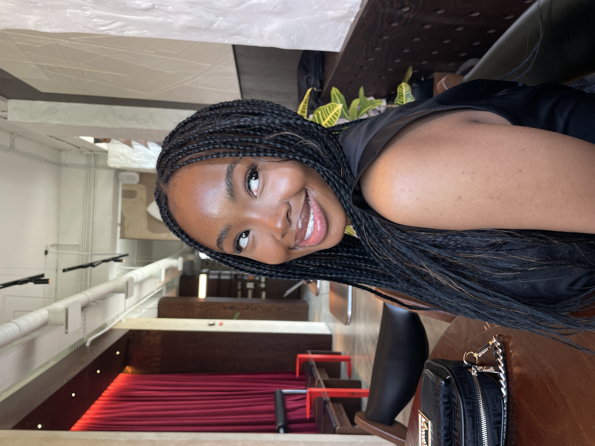 A medium shot captures a young Black woman with braided hair looking over her left shoulder and smiling into the camera. She is seated at a wooden table with a black purse on it, and appears to be in an indoor setting with a modern, industrial aesthetic. Red velvet curtains hang in the background, and there are exposed pipes and lighting fixtures on the ceiling. The woman is wearing a black sleeveless top, and her expression is bright and engaging.