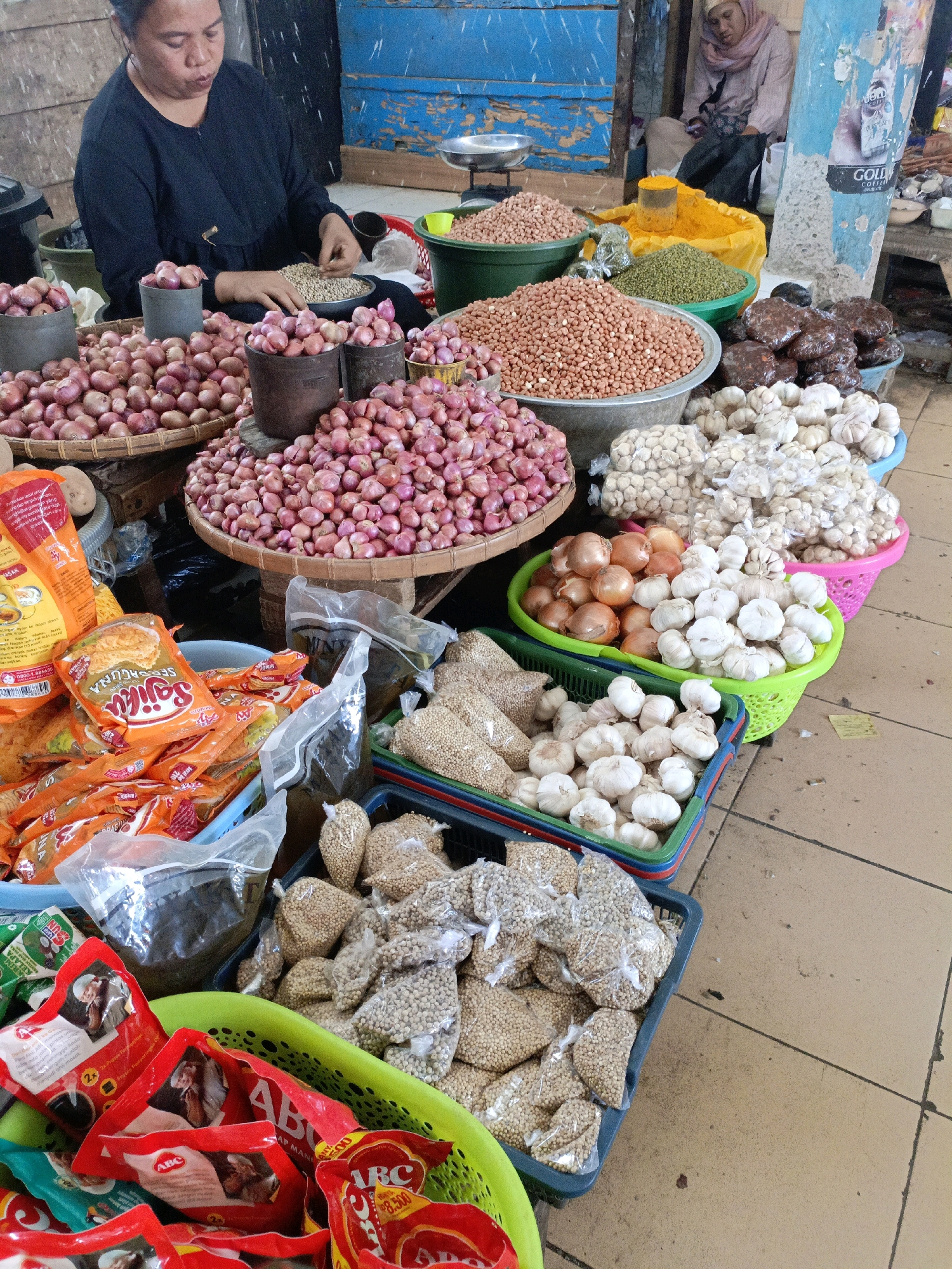 The image depicts a bustling market scene in Bulukumba, Indonesia, with a focus on various food items being sold. In the foreground, a woman is seated, sorting through small white grains. Surrounding her are large piles of red onions, some displayed in woven baskets and metal containers. To the right, there are several colorful plastic baskets filled with produce and dried goods. One basket holds garlic bulbs, another contains brown onions, and further to the right, more baskets are filled with various legumes and spices, including what appears to be peanuts and green beans. Packaged food items, predominantly in orange and red wrappers, are also visible in a blue basket in the lower left corner. In the background, another woman, wearing a headscarf, is seated, appearing to rest. The market stalls are constructed from wood and metal, with blue and beige colored walls visible. The floor is tiled, and the overall impression is one of an active and diverse marketplace.