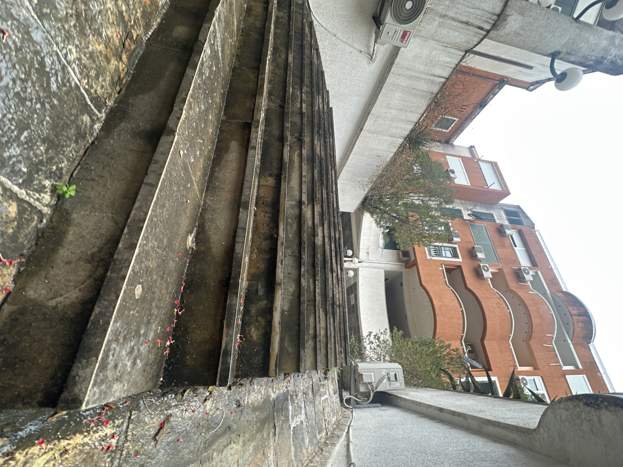 The photograph captures a low-angle perspective of a building and its surroundings in Budva, Montenegro. In the foreground, concrete stairs with a vertical ridged design are visible. Scattered red debris lies on the stairs and along the base of the structure. Above the stairs, a building with a white exterior houses an air conditioning unit. In the background, a taller building with a curved, orange facade and numerous windows and air conditioning units is prominent. The sky appears overcast, and various trees and bushes are scattered throughout the scene, adding to the urban environment.
