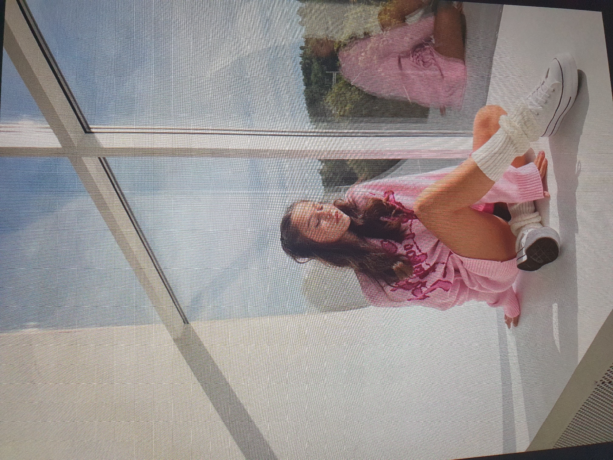 A young woman with long, dark hair is lying down on a white surface, positioned near a large window. She is wearing a pink, long-sleeved top with a graphic design and pink knit leg warmers over white sneakers. Her eyes are closed, and she appears relaxed. The scene is brightly lit, suggesting it is daytime. The large window reflects the sky and some greenery, indicating an outdoor view. The overall aesthetic is casual and bright, possibly a photoshoot or a moment of leisure. The woman's pose and attire suggest a relaxed and comfortable atmosphere.