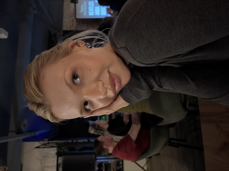 The scene depicts an indoor establishment, likely a bar or restaurant, located within the city of Chaikovsky, Russia. The specific venue name or address cannot be determined from the image.
The primary subject is a light-skinned female with blonde hair pulled back, visible from the chest up, wearing a dark grey long-sleeved top. She props her left hand against her cheek and gazes upward and to her right with a gentle expression. A small metallic labret piercing adorns her lower lip, and she has a silver hoop earring in her left ear, along with another small cartilage piercing.
In the blurred background, several patrons are seated at tables. To the left, a man in a red shirt is observed from behind, seated near a dark speaker. Further back, another person with braided hair is visible. The environment features an exposed brick wall, dark industrial-style ceiling fixtures, and blue ambient lighting. On the right, a large projection screen displays musical artist names including "BEATLES," "ABBA," and "QUEEN," suggesting a themed event or live music performance. A dark wooden table surface occupies the lower foreground.