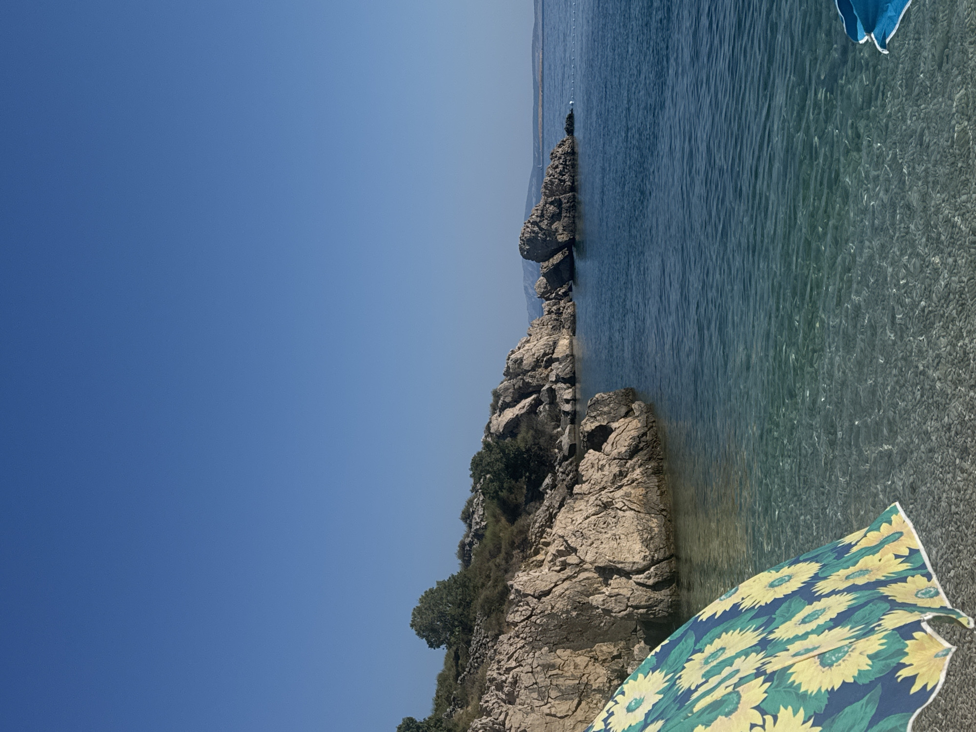 The image depicts a rocky coastline and a clear, calm body of water, likely the Adriatic Sea, with a bright blue sky overhead. In the foreground, a portion of a beach umbrella with a sunflower pattern is visible on a pebble beach. The water near the shore is shallow and transparent, revealing the pebbled seabed. Further out, the water deepens to a darker blue. Rocky formations rise from the sea and along the coastline, with sparse vegetation dotting the landscape. In the distance, a hazy landmass or island is visible on the horizon. The scene conveys a tranquil atmosphere typical of a summer day at a coastal resort.
