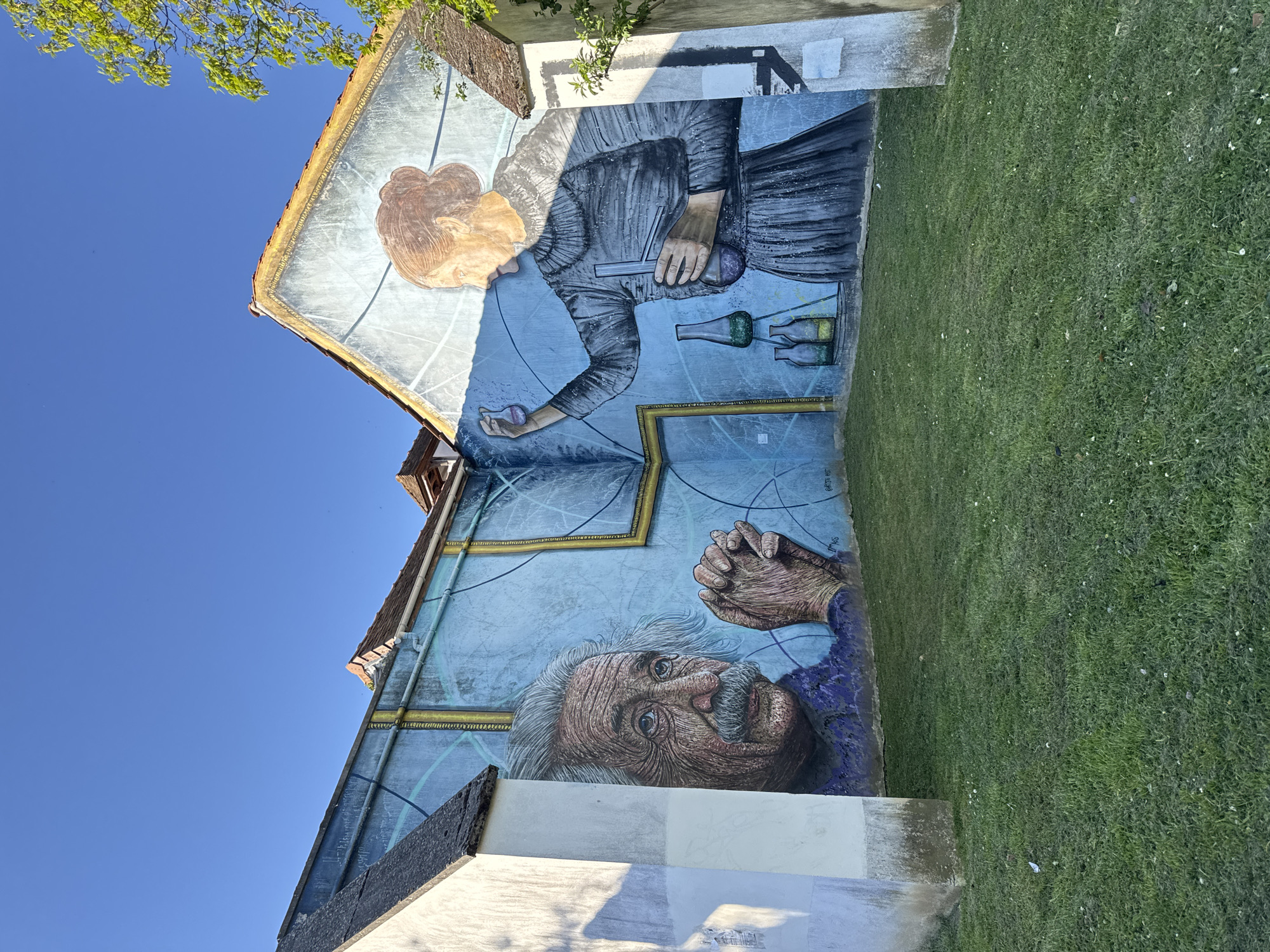 The image captures a large mural on the exterior of a building in Palaiseau, France. The mural depicts two historical figures: Marie Curie and Albert Einstein. Marie Curie is illustrated in a monochromatic style, holding a test tube and what appears to be a bulb, possibly referencing her work with radioactivity. Several glass bottles are also included in the scene. Below Marie Curie, Einstein is depicted, with his hands folded in front of him. The mural is painted in shades of blue and features a background with what appears to be circular, scientific or mathematical diagrams. The building is situated next to a green lawn and the sky is a clear blue.