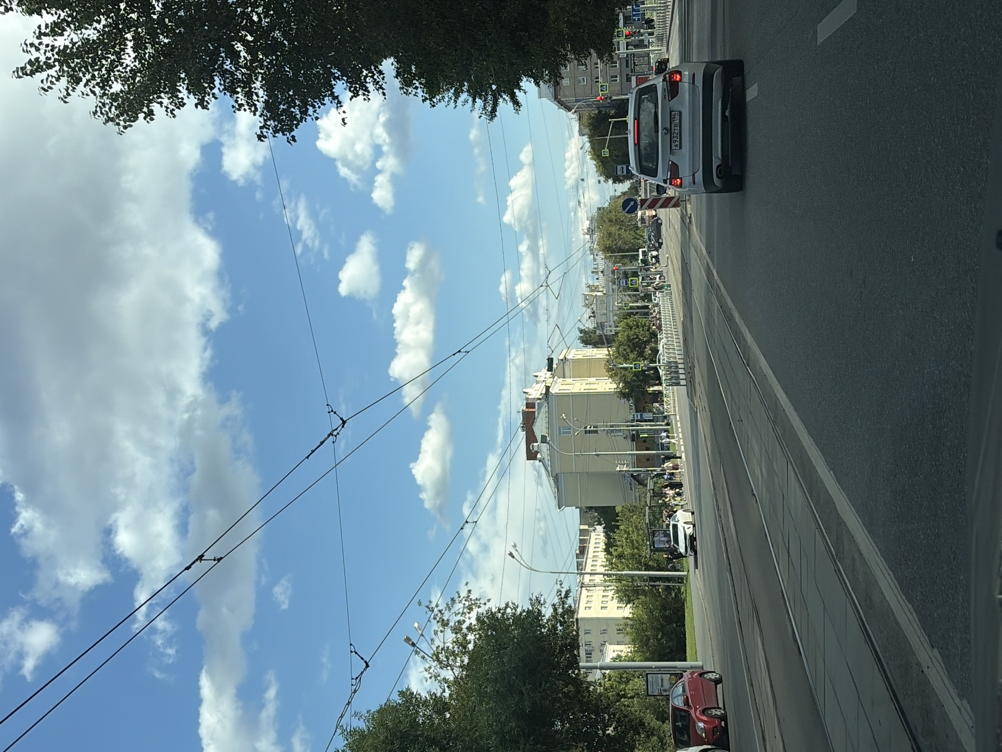 The image depicts a street scene in Yekaterinburg, Russia, under a sky with scattered clouds. A multi-lane road is visible, with what appear to be tram tracks running along one side. Several cars are on the road, including a white BMW SUV in the foreground. Buildings line the street, with trees and overhead wires, likely for public transportation, also present. Pedestrians can be seen near a bus stop. The overall environment suggests a moderately busy urban setting during daylight.