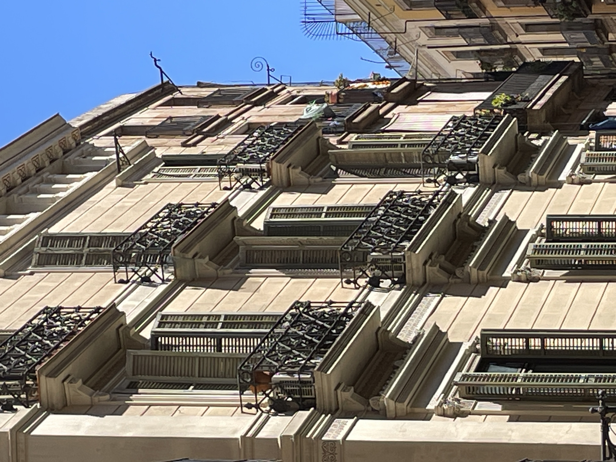 This image captures a close-up, low-angle view of an ornate building facade in Barcelona, Spain, under a clear, bright blue sky. The building, constructed from light-colored stone or stucco, exhibits multiple stories with a repetitive pattern of windows and balconies.
Each balcony is adorned with intricate black wrought-iron railings featuring detailed, geometric patterns. Many of these balconies are recessed, forming deep alcoves with architectural details like cornices and decorative brackets. Some balconies also appear to have small planters or greenery, particularly on the upper levels. The windows behind the railings are partially visible, with some suggesting open shutters or internal blinds.
The upper portion of the building and its roofline reveal various metallic structures, including what appear to be antennae or decorative ironwork, one of which has a distinct spiral shape. Small patches of green foliage are scattered along the roof and upper ledges.
The scene is illuminated by strong, direct sunlight, creating sharp shadows that accentuate the three-dimensional depth and architectural details of the facade. The weather is clear and sunny, with no clouds in the bright blue sky. There are no people visible, and no specific activities are taking place, allowing the focus to remain on the architectural beauty and detailing of the building. No visible text is present in the image.