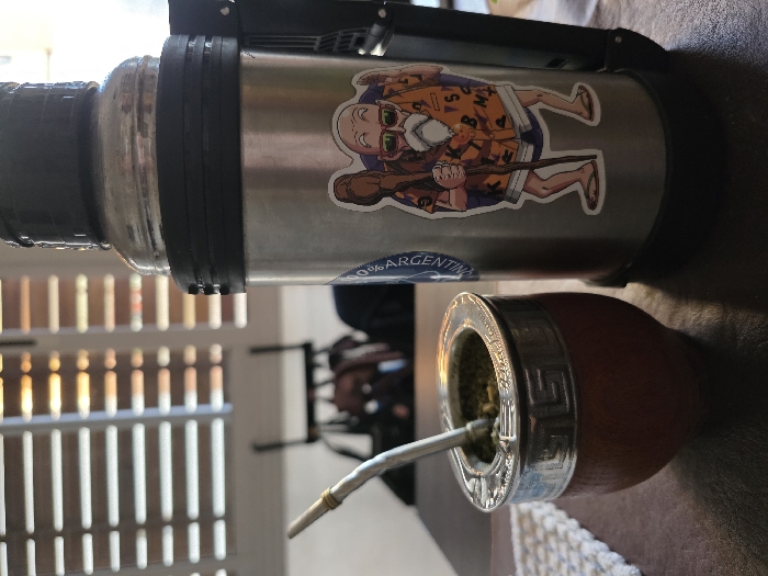 This image captures an indoor scene in Villa San Andrés, Argentina, featuring a traditional mate setup. In the foreground, a brown calabash mate gourd with a polished silver-colored metal rim, decorated with a repeating geometric pattern, sits on a dark, flat surface. The gourd contains green yerba mate leaves, with a silver bombilla (straw) inserted. To the immediate right, a tall, cylindrical stainless steel thermos stands upright. This thermos has a black plastic base and an integrated black handle/pour mechanism on its right side; its upper section consists of a clear, possibly glass or plastic, liner. A partially obscured circular blue sticker is visible on the left side of the thermos, and a prominent sticker depicting an animated elderly male character with a white beard, sunglasses, an orange Hawaiian-style shirt, shorts, and flip-flops, holding a staff, is affixed to the right side of the thermos. A white, knitted or crocheted mat is partially visible beneath the mate gourd on the left. The background is blurred, showing a window with white horizontal louvered blinds through which sunlight filters, creating distinct horizontal strips of light and shadow. Indistinct dark shapes of furniture or other objects are visible further in the background.