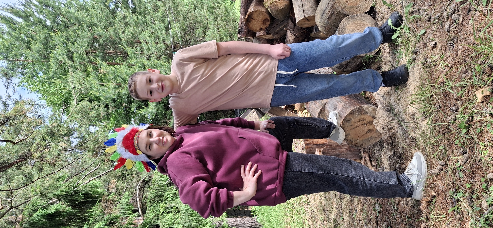 Two people are in an outdoor setting near Прикамский, Russia. One is a person wearing a maroon hoodie and a colorful headdress; the other is a child wearing a tan shirt and jeans. Both individuals are positioned on or near logs. The background includes trees and foliage, indicating a wooded or park-like environment.