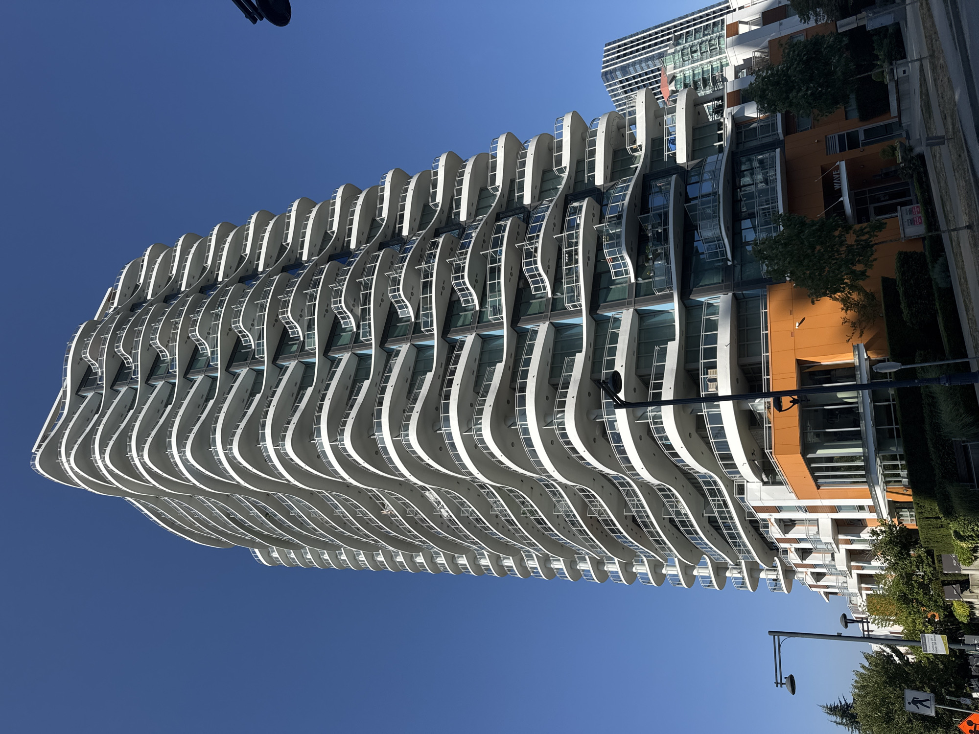 The image captures a modern, curved high-rise apartment building with numerous balconies, set against a clear blue sky. The building features a distinctive white, undulating facade with glass railings on the balconies. The lower levels of the building are clad in orange panels and feature large windows, suggesting commercial or residential spaces. A street scene is visible at the bottom of the image, with sidewalks, trees, streetlights, and traffic signs. Another taller, angular building can be seen in the background to the right. The presence of signage, including a street sign and a sign reading "WAVE," indicates an urban environment. The overall impression is a contemporary architectural design within a city setting.
