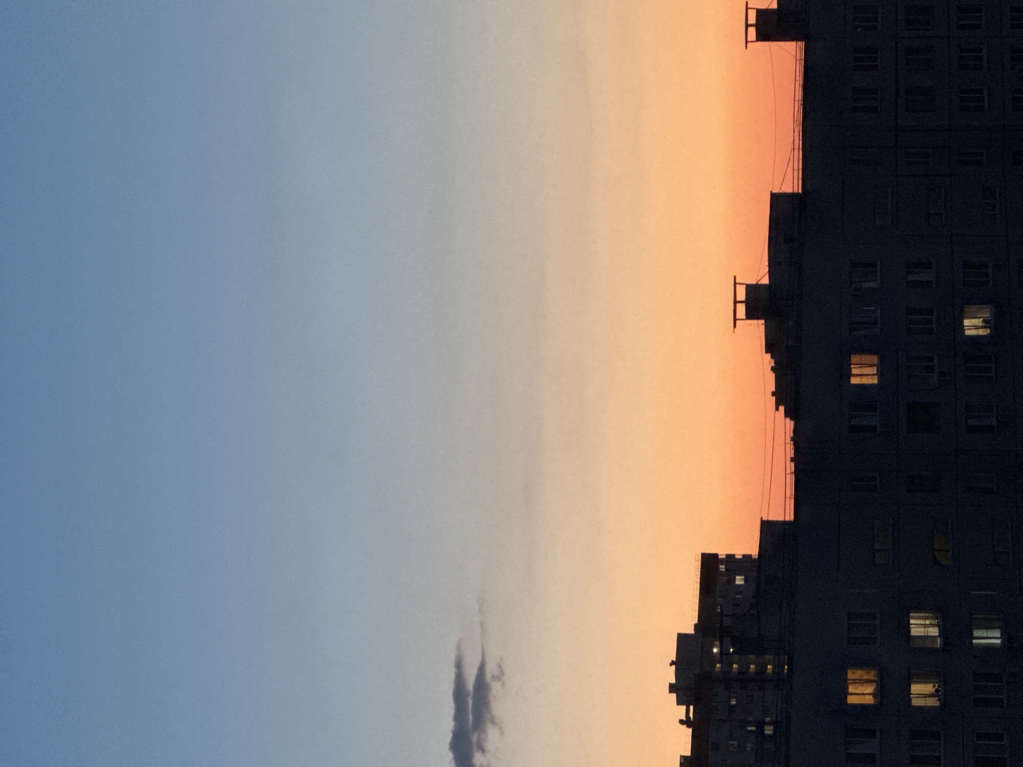This is a vertical, full-frame shot of a city skyline at dusk. The sky transitions from a pale blue at the top to a vibrant orange at the horizon. A wispy, dark gray cloud is visible in the upper left quadrant. Silhouetted against the sunset are multiple apartment buildings. The closest building in the foreground is dark gray and shows many windows, some of which are illuminated with a warm yellow light. Behind this building are two taller structures, also silhouetted and dark. These buildings have distinct geometric shapes on their rooftops, resembling chimneys or ventilation shafts. The overall impression is that of a residential urban environment at the end of the day.
