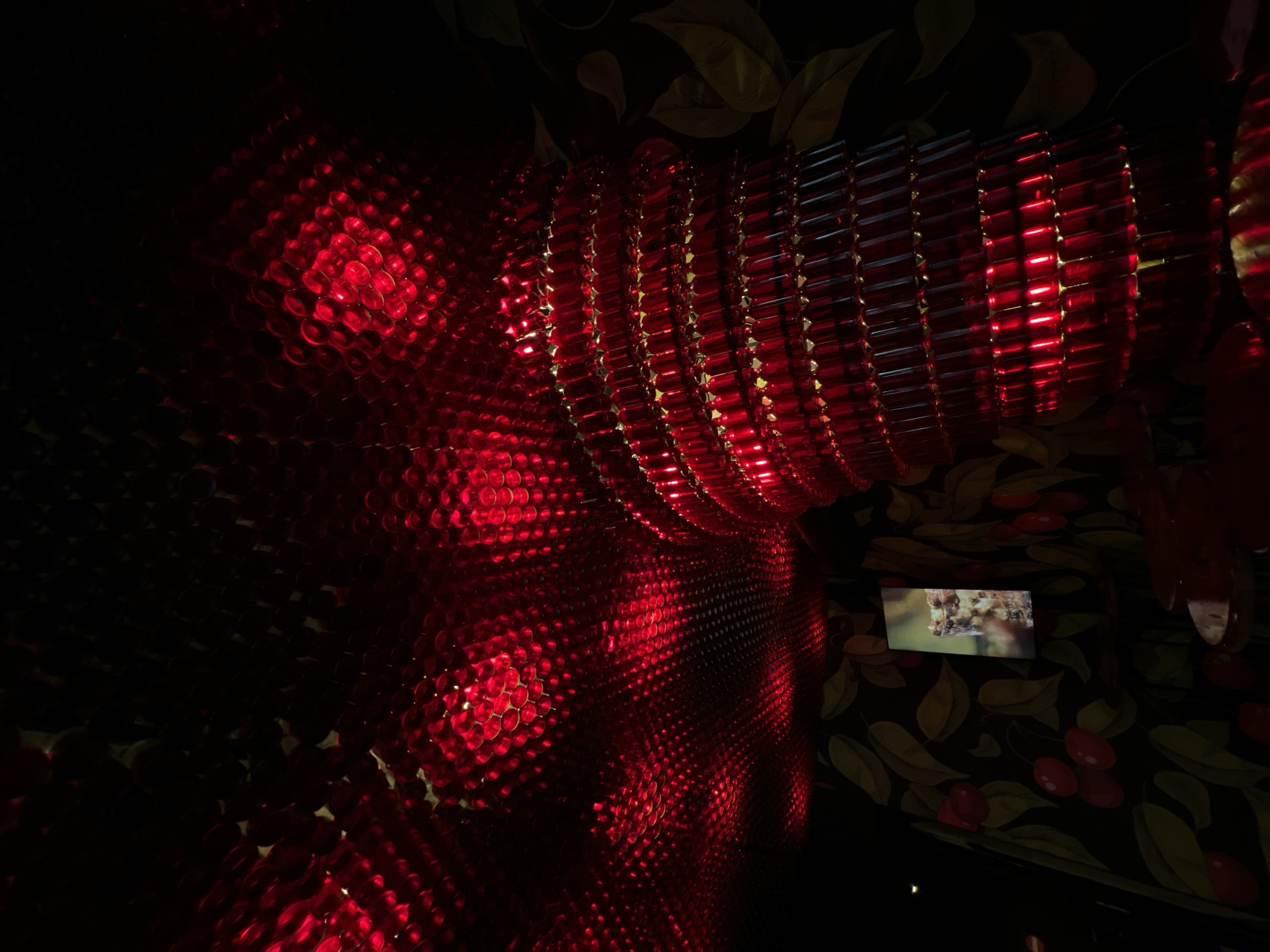 The image depicts an interior space, possibly a restaurant or bar, illuminated by red lighting. The ceiling is adorned with a textured surface reflecting the red light, creating a patterned effect. A large, cylindrical structure, seemingly made of stacked red bottles or glass elements, is prominently featured in the foreground. The walls are covered in dark wallpaper with a repeating pattern of green leaves and red cherries. A small screen displaying an image of food or a beverage is mounted on one of the walls. The overall ambiance is dark and intimate, with the red lighting creating a dramatic and perhaps festive atmosphere. The location of this scene is not specified in the image itself and cannot be determined from the visual information provided. Therefore, it is not possible to confirm or deny if it is located in the city of Lublin, Poland.