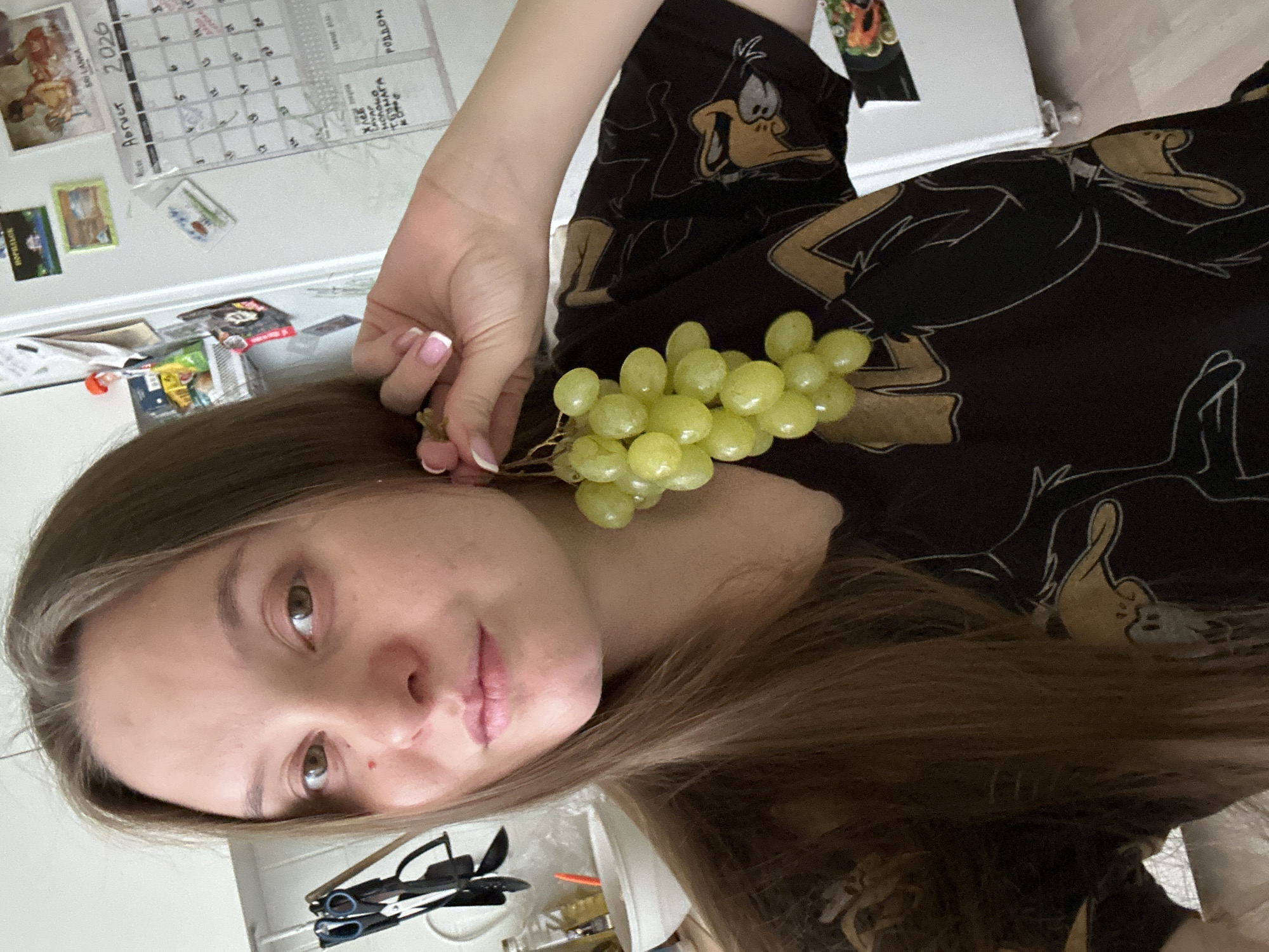 A woman with long brown hair is holding a bunch of green grapes to her ear as if it were an earring. She is wearing a black t-shirt with a cartoon pattern. Behind her, a white refrigerator door is visible, adorned with magnets and a calendar showing "Август 2026" (August 2026). There is also a rack with kitchen utensils and a bowl on a counter. The scene appears to be a kitchen, likely in a residential setting. The mention of Noviy, Russia is not verifiable from the image itself.