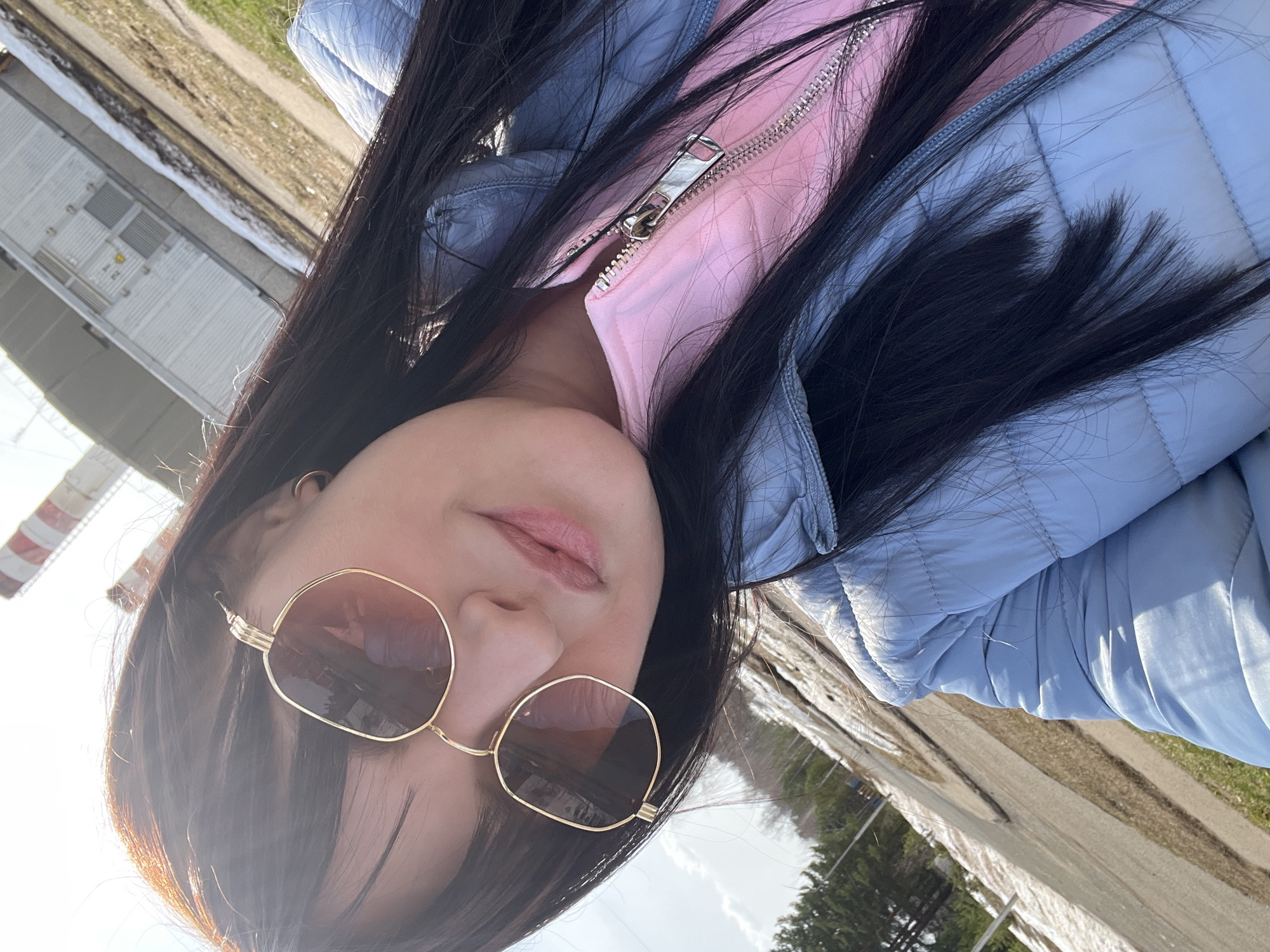 A woman with long dark hair and sunglasses is pictured in a selfie. She is wearing a light blue puffer jacket over a pink top. The background includes a road, grass, trees, a partially snow-covered area, and a tall structure with red and white stripes. The location is likely in the city of Марковский, Russia.