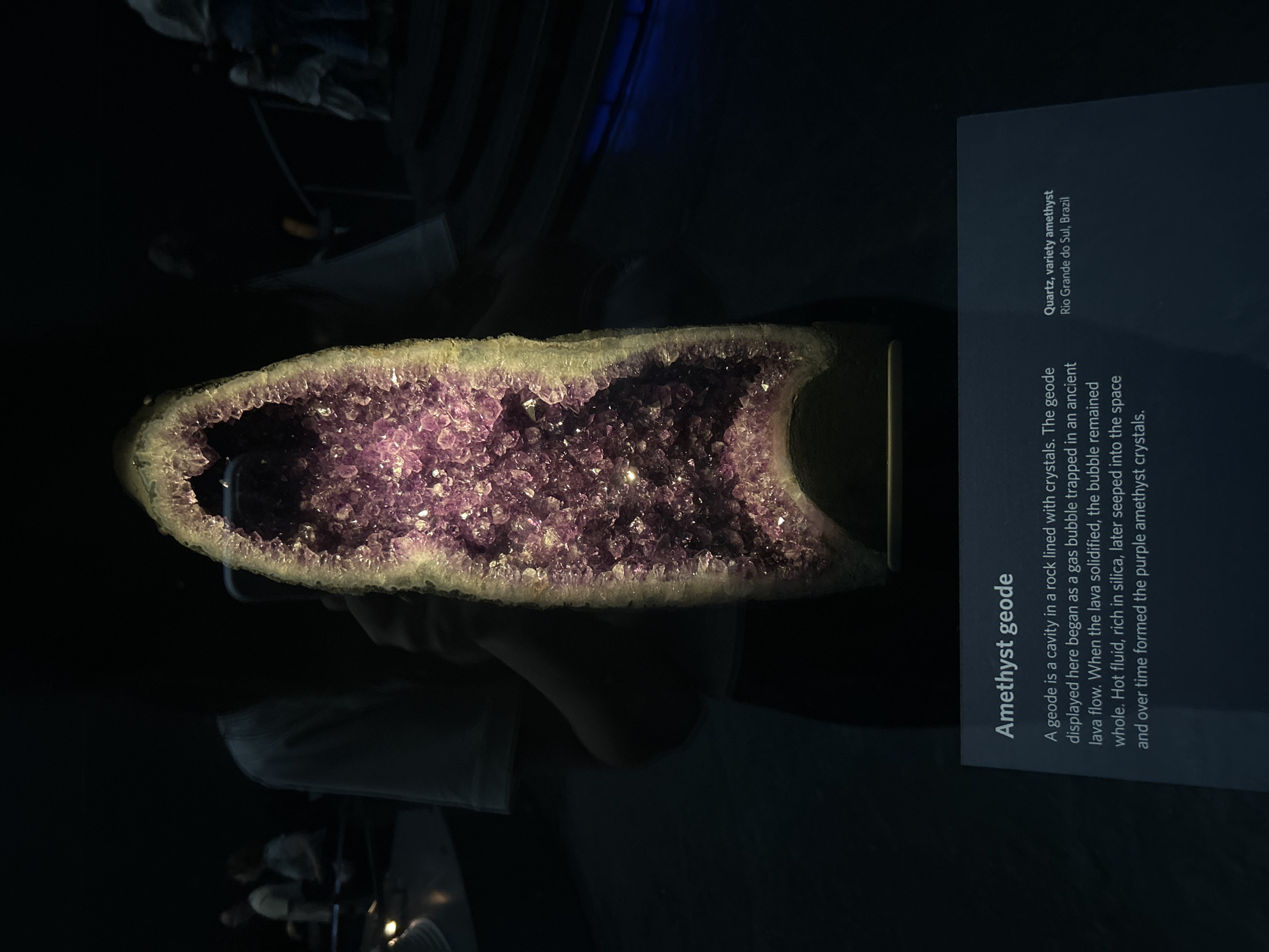The image shows a large amethyst geode on display in a dimly lit environment, likely a museum. The geode is prominent in the center, its interior cavity filled with sparkling purple crystals. The outer edge of the geode is rough and a lighter, yellowish-brown color. A dark base supports the geode. To the left of the geode, partially obscured by the geode itself, is a sign with text. The sign's title is "Amethyst geode" followed by a description of its formation. To the right of the geode and slightly lower is another section of the sign with the text "Quartz, variety amethyst Rio Grande do Sul, Brazil". In the background, out of focus and in shadow, are several people. One person is holding up a smartphone, possibly taking a picture of the geode. Another person is visible in the upper left, and further back, more people are seen on elevated platforms or stairs, suggesting a public exhibition space. The overall ambiance is dark, with focused lighting on the geode. The exact location in London, United Kingdom cannot be determined from the image.