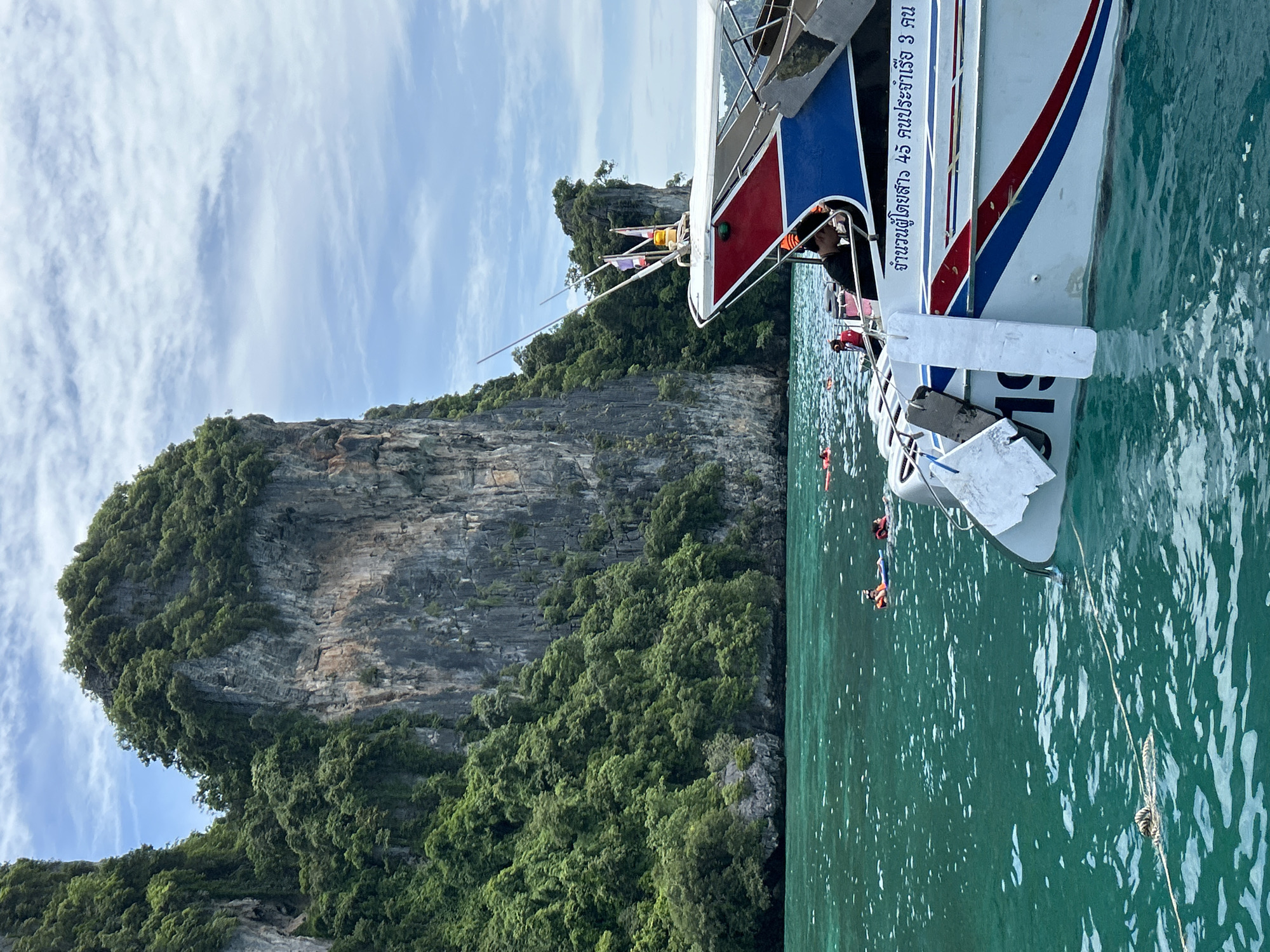 The image depicts a scene in Ao Nang, Thailand, featuring a speed boat partially submerged in clear turquoise water. The boat has a red, white, and blue design with Thai writing on its side. Several people are visible in the water, some wearing orange life vests and snorkeling gear, suggesting they are engaged in water activities. In the background, towering limestone cliffs covered in lush green vegetation rise dramatically from the sea under a partially cloudy blue sky. The overall environment suggests a popular tourist destination known for its natural beauty and water-based recreation.