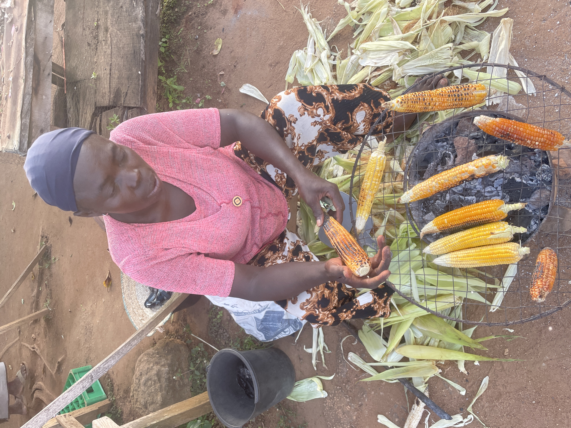 A woman, wearing a pink top and a black and white patterned skirt, is grilling corn over charcoal. She is sitting on the ground, with a collection of corn husks scattered around her. The corn is arranged on a metal grill, with some ears already roasted to a golden brown and others still being cooked. The woman is holding a freshly grilled ear of corn in her hand, examining it. The scene appears to be outdoors, with a dirt ground and some wooden structures in the background. The lighting suggests it is daytime. The overall atmosphere is one of food preparation in a rustic setting. There is no visible text in the image.