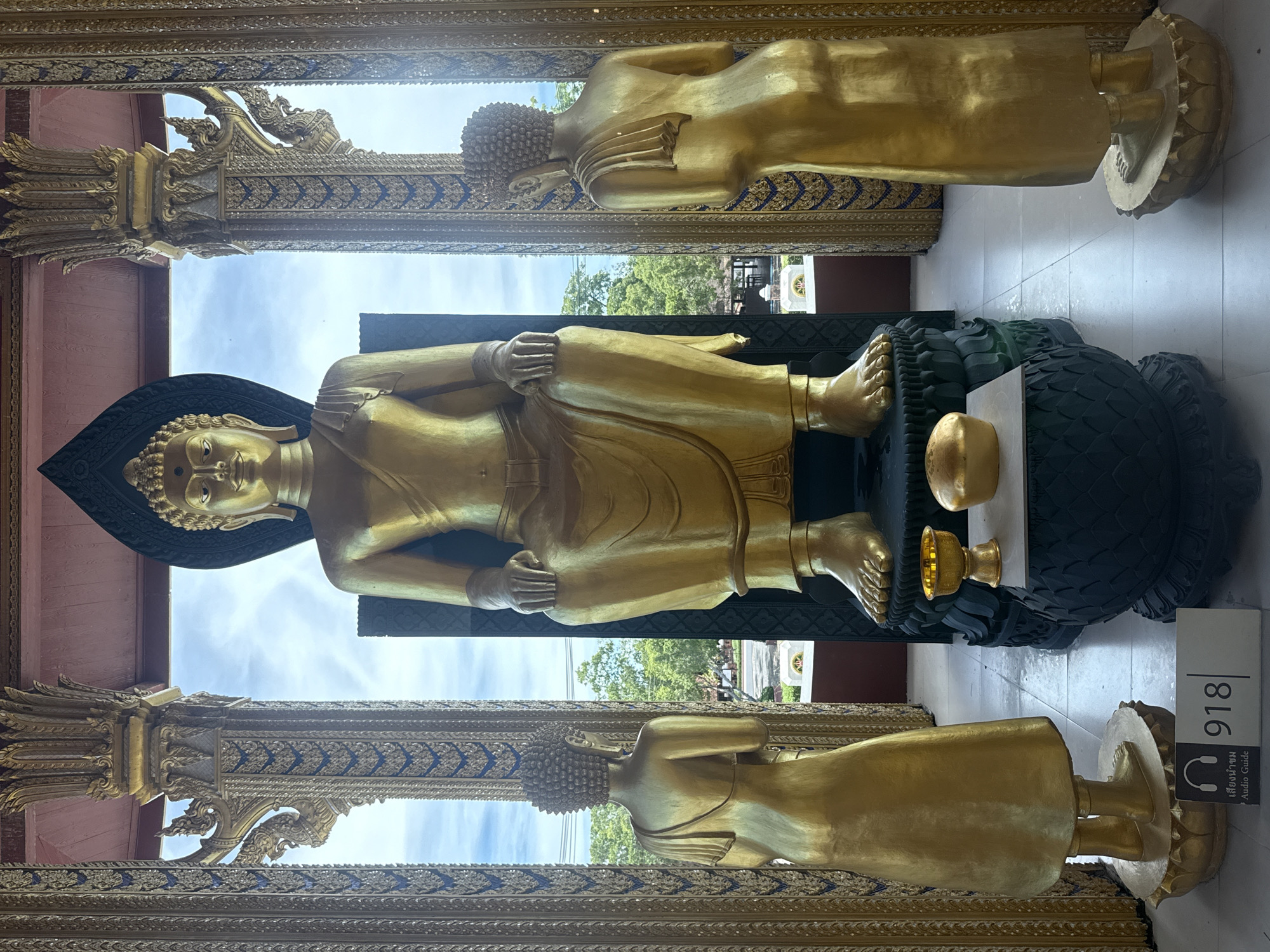 A horizontal eye-level shot shows a golden reclining Buddha statue in Bang Pu Subdistrict Municipality, Thailand. The statue is flanked by two similar standing golden statues at the top and bottom of the frame.
The reclining Buddha, the main subject, lies horizontally across the center of the frame, facing forward. It is golden, with a black headrest. Its hands are placed on its upper torso. A golden offering sits beside the statue’s feet on a dark base that resembles snake scales.
Two additional golden statues stand, one above and one below the reclining Buddha. They face away from the viewer. The top portion of the frame shows an ornate gold and blue structure, mirroring a similar structure at the bottom of the frame. The area visible between these structures shows a view of trees and sky, indicating an outdoor setting. To the right of the reclining Buddha’s feet is a sign with the number “918” and an icon of headphones, suggesting an audio guide. The floor is a light-colored tile.