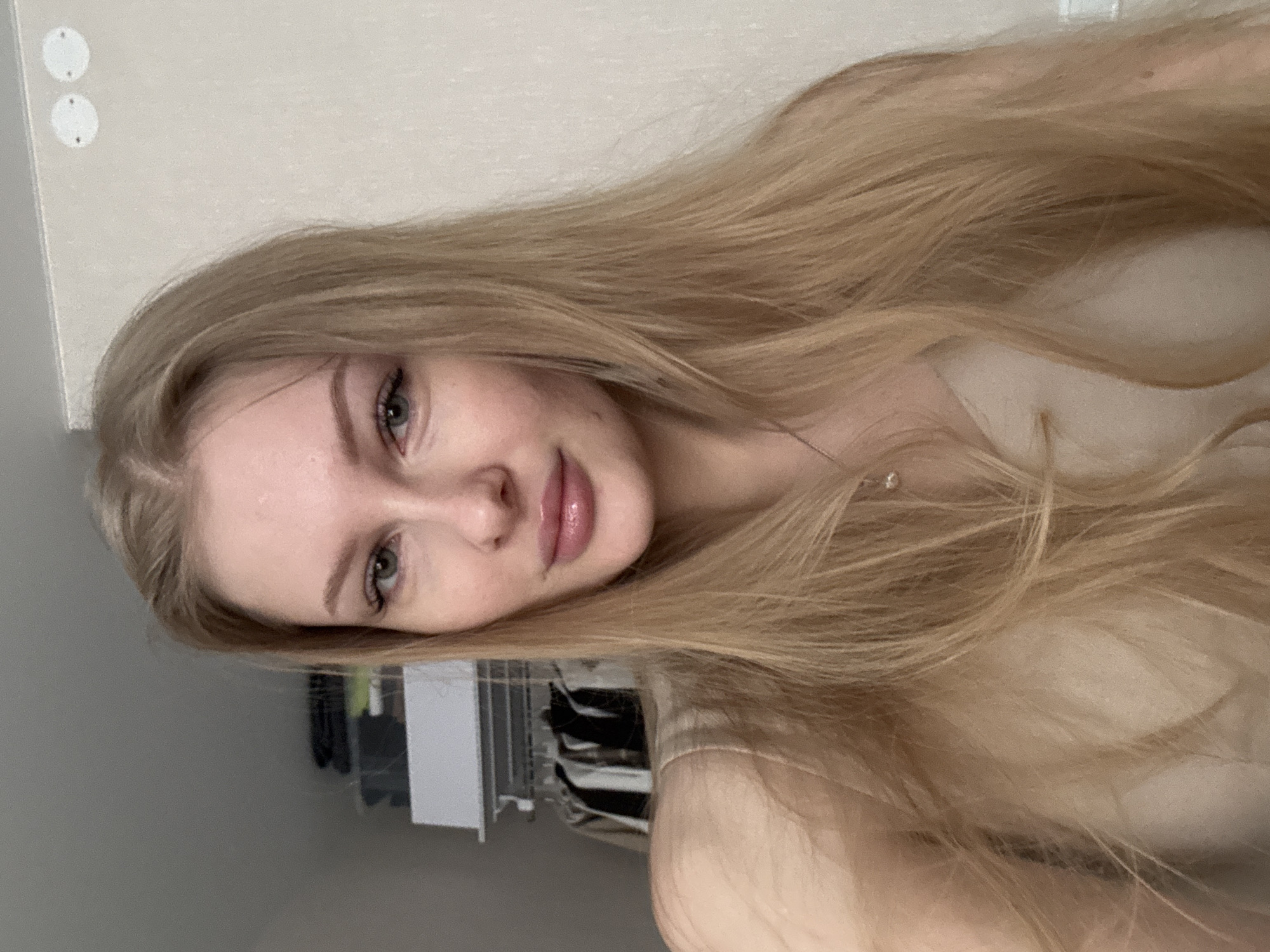 A close-up selfie of a young woman with long, light blonde hair and green eyes. She has fair skin and is wearing what appears to be a beige top. The background shows a wall with light-colored paint and a glimpse of a clothing rack. There is no clear indication of the city of Perm, Russia, or any specific location.