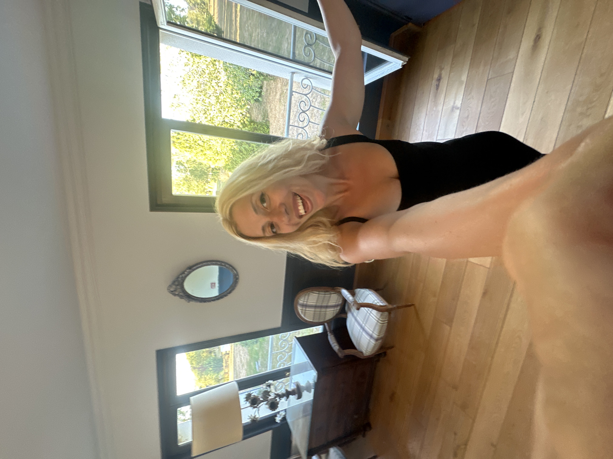 A woman with blonde hair and a black dress takes a selfie in a room with wooden floors and large windows. She is smiling and looking at the camera. The room contains a dark wooden dresser with a lamp and a decorative item on top, and a small armchair with a plaid fabric. Outside the windows, trees and greenery are visible. The image is taken from a low angle, capturing the woman's arm and hand in the foreground. The location is identified as Zutkerque, France.