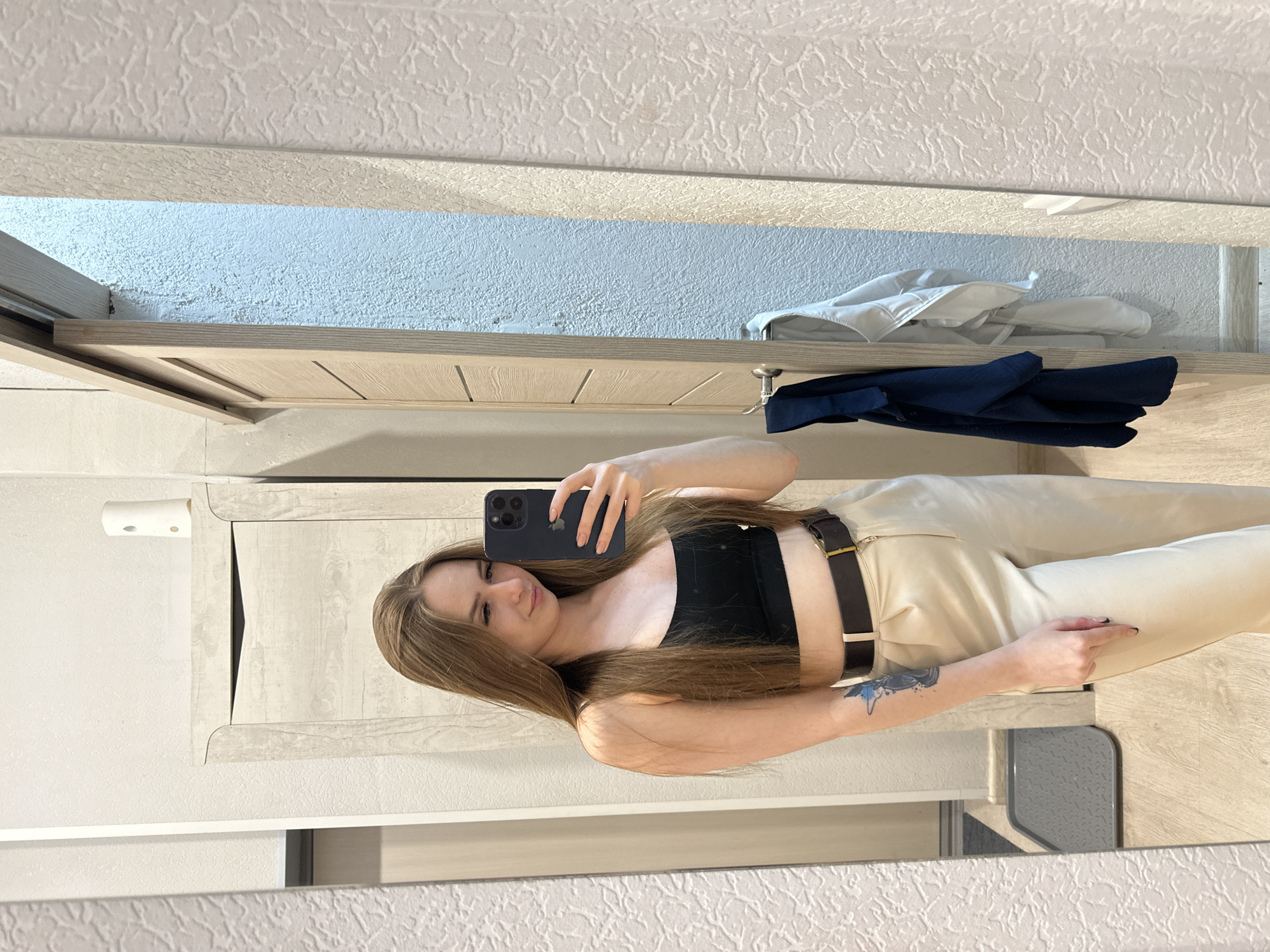 The image shows a woman taking a selfie in front of a mirror. She is wearing a black top, beige pants, and a dark belt. Her hair is long and brown. There is a cabinet and some clothes visible in the background, and the room appears to be indoors. The location is Unknown, Unknown.
